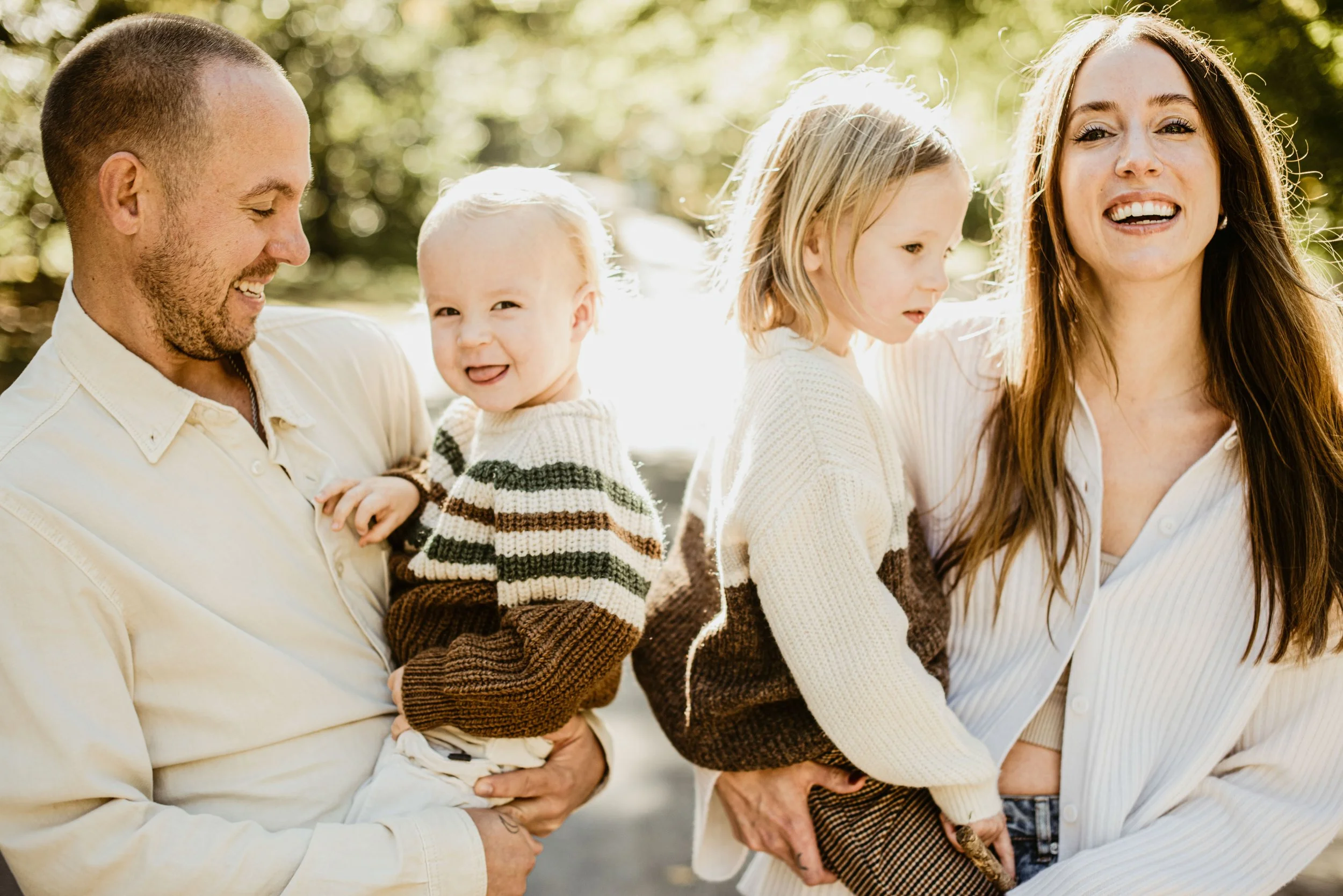 A family of four enjoying outdoors on a sunny day. The father on the left is smiling while holding a young child. The mother on the right is smiling and holding a small girl, with trees and sunlight in the background.