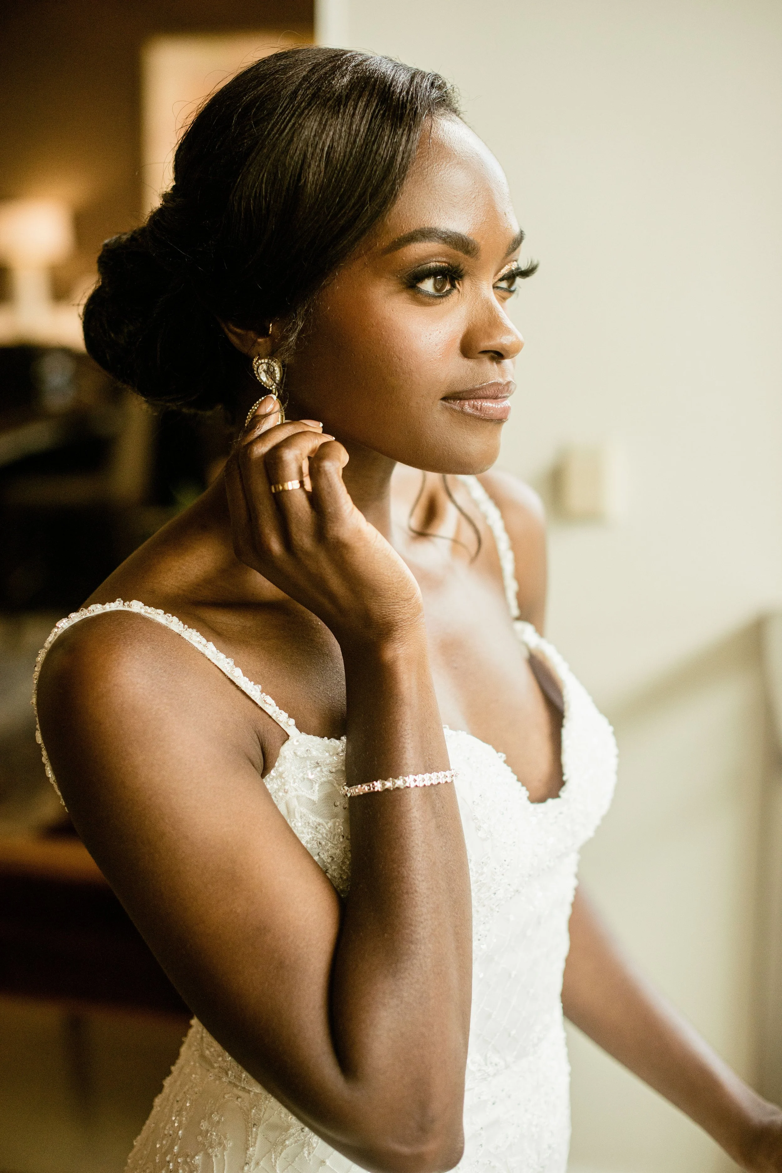A woman with dark skin and long dark hair styled in an updo, wearing a white lace dress and jewelry, looking to the right, close-up profile shot in a softly lit room.