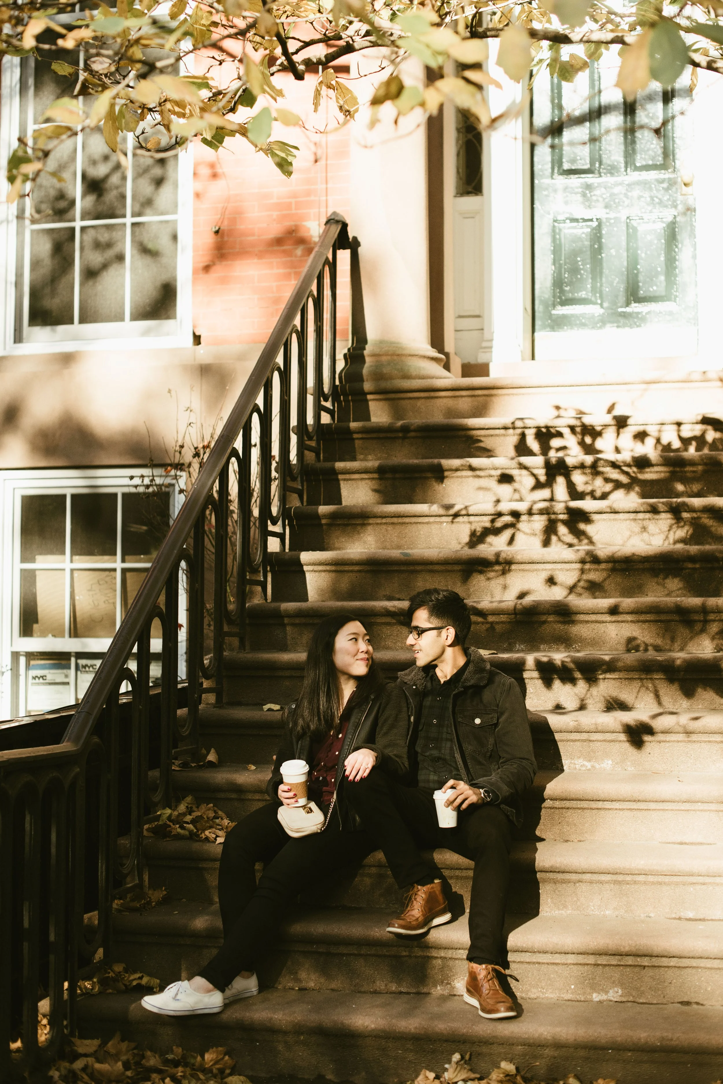 A young man and woman sitting on outdoor stairs, holding coffee cups, smiling and looking at each other.