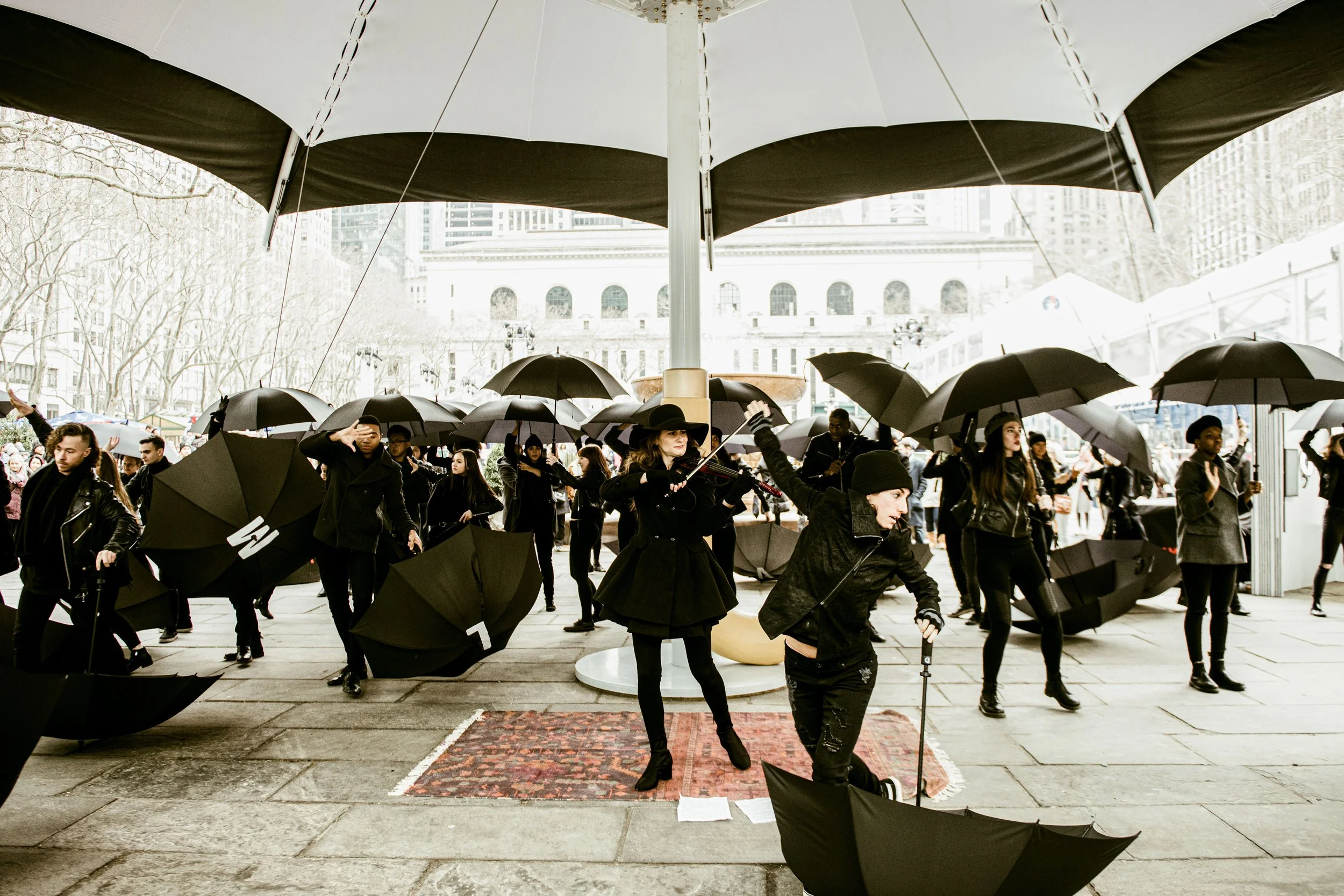 A group of people participating in a street event or performance, holding black umbrellas, with some conducting or playing musical instruments, under a large white and black umbrella structure in an urban area.