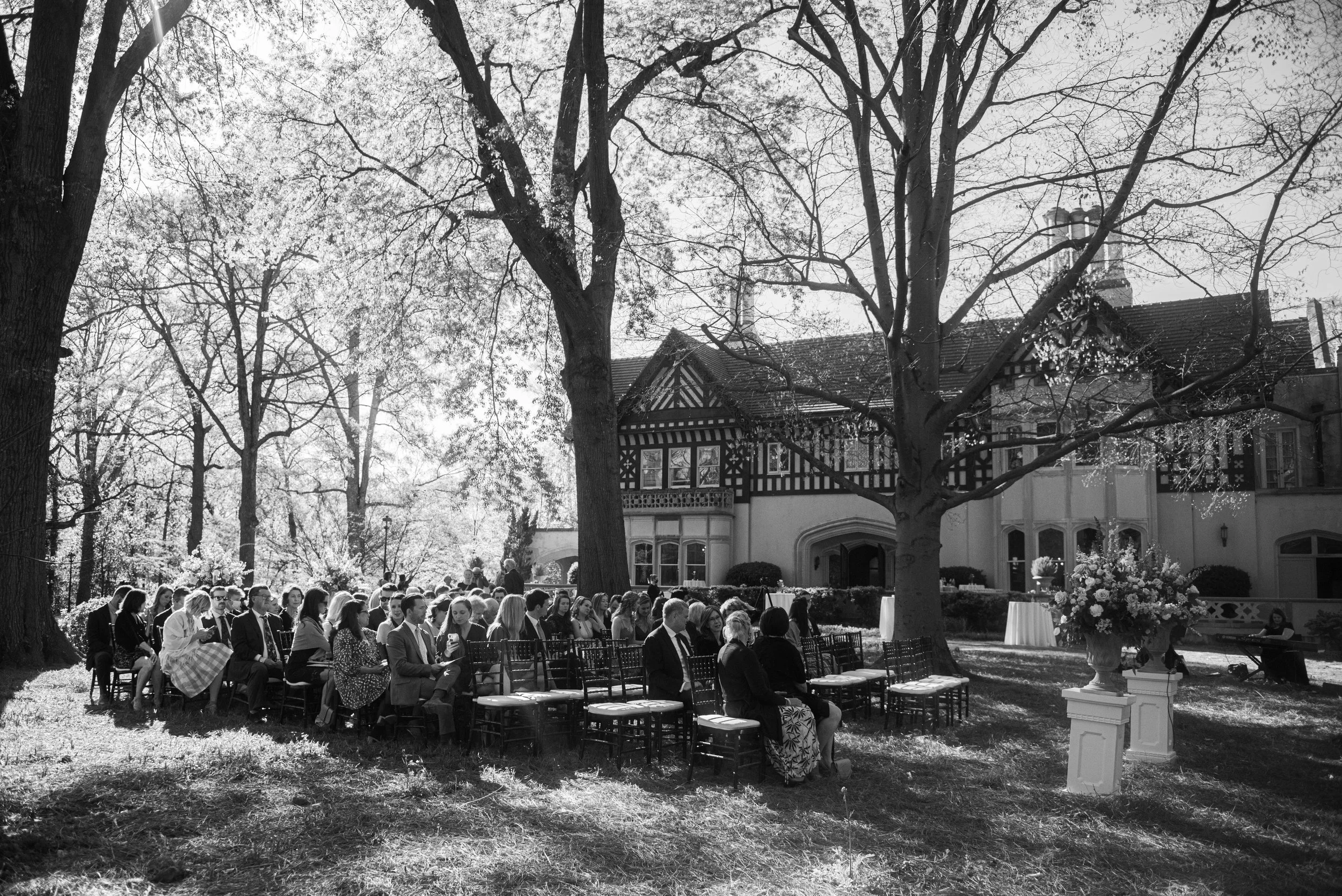 Black and white photo of an outdoor formal gathering with rows of seated people under large trees in front of a historic estate or mansion, with floral arrangements and a cake table.