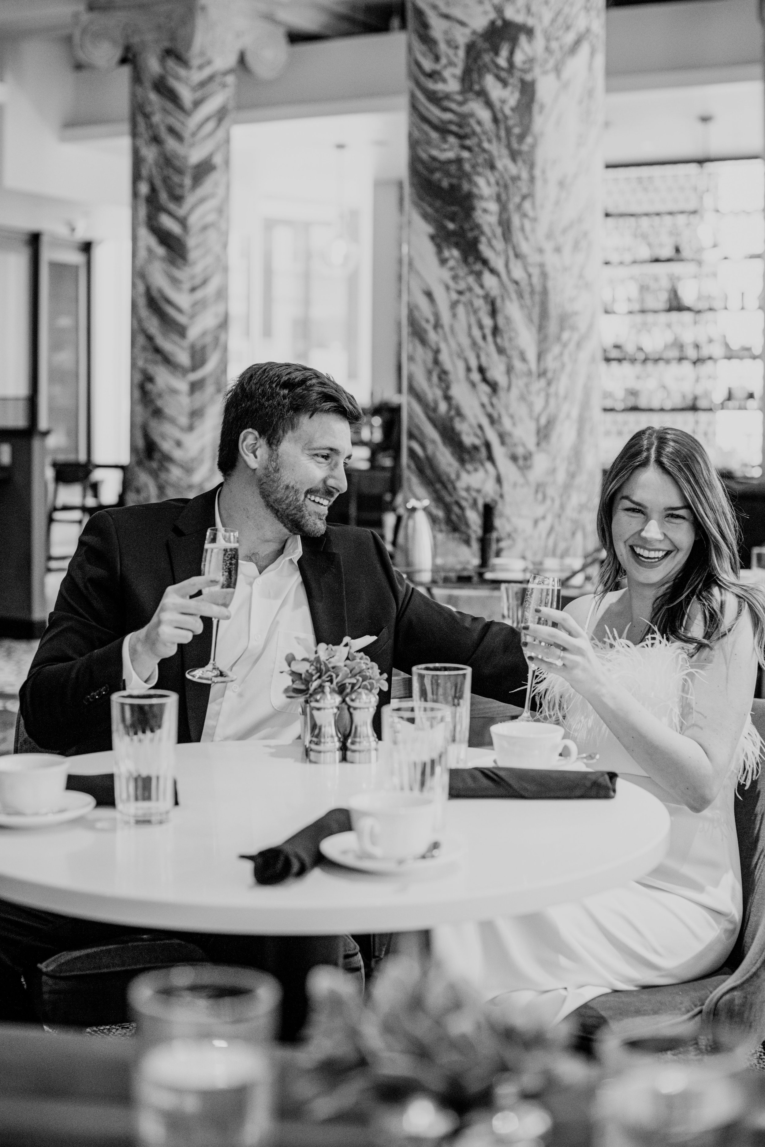 A man and woman sitting at a dining table in a restaurant, smiling and toasting with glasses of wine. The man has a beard, wearing a suit jacket and white shirt. The woman has long hair, wearing a dress with feather-like details. The table has a flor