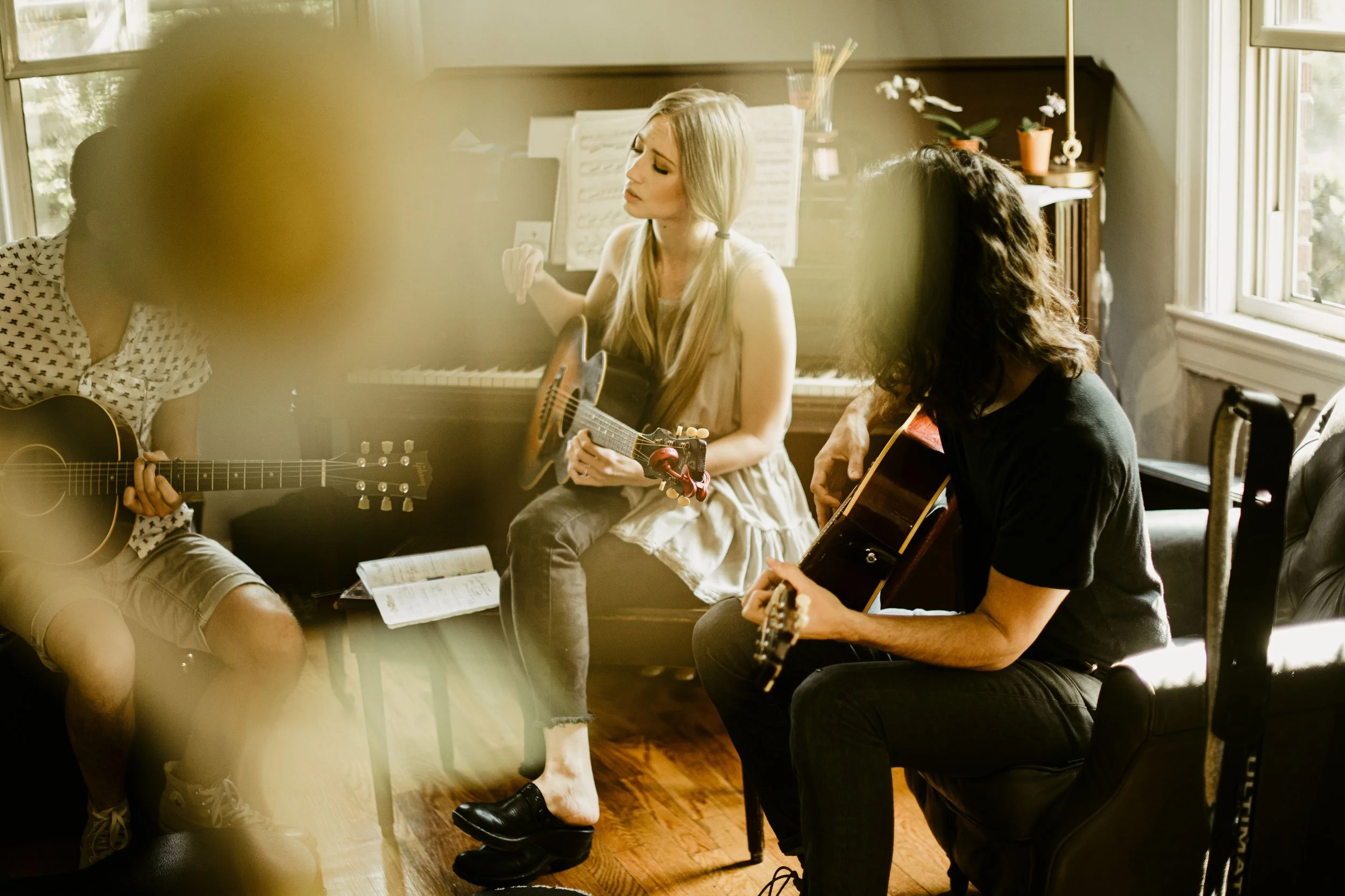 Three women sitting in a cozy room, playing guitars and singing, with music sheets on a piano behind them and sunlight streaming through the window.