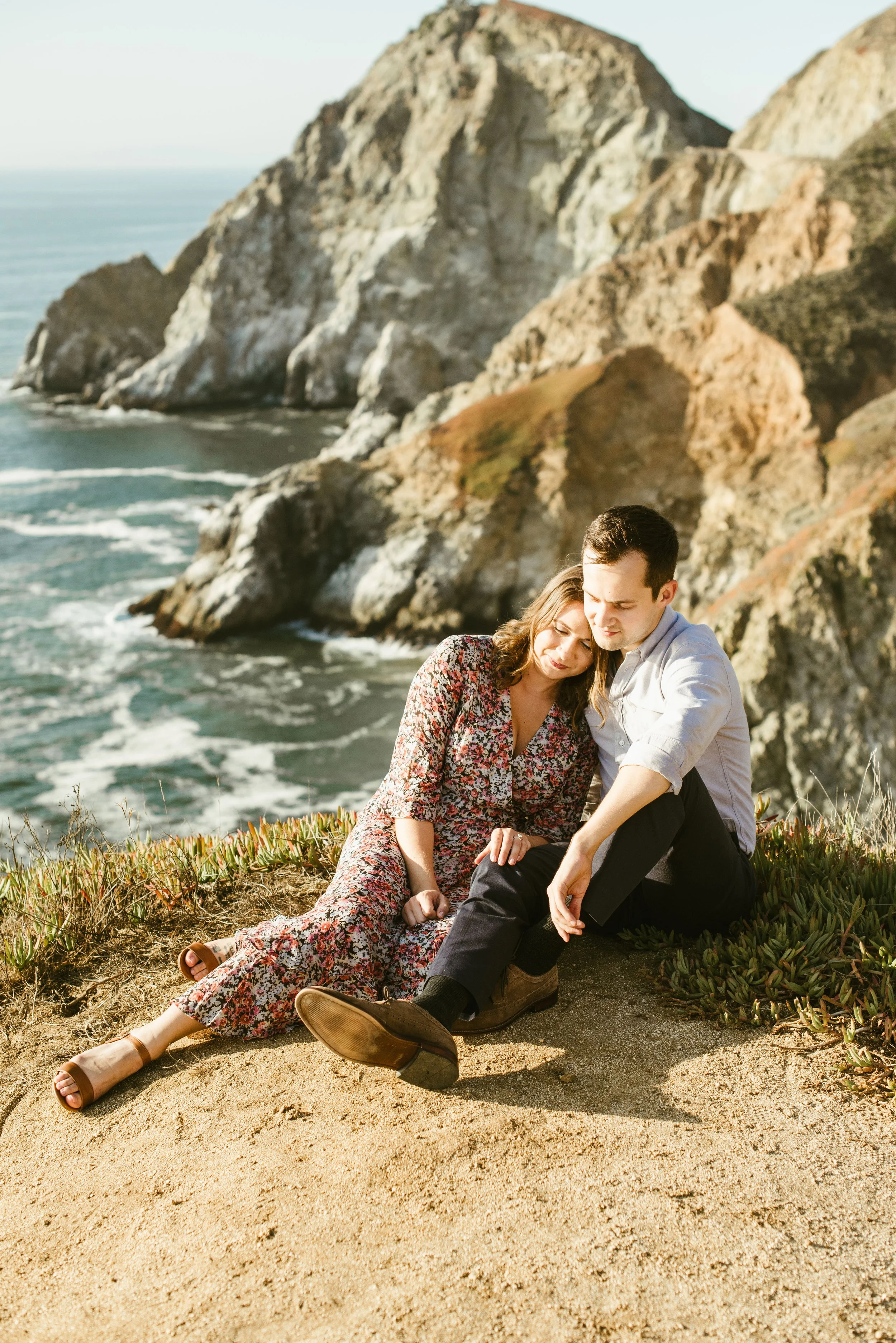 A couple sitting on a sandy beach with cliffs and the ocean in the background.