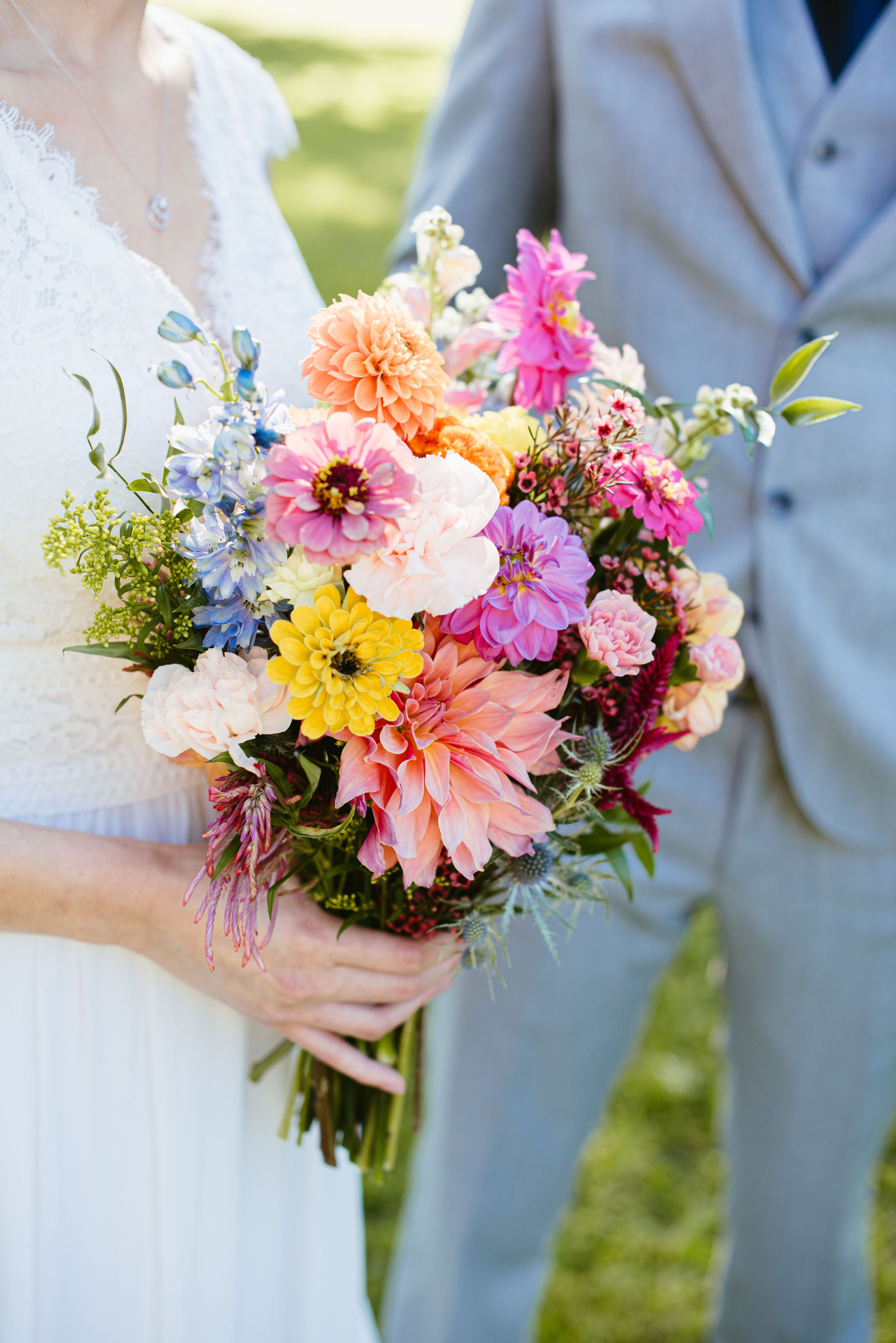 A person in a white dress holding a colorful bouquet of flowers, with another person in a light gray suit beside them, outdoors in a sunny setting.