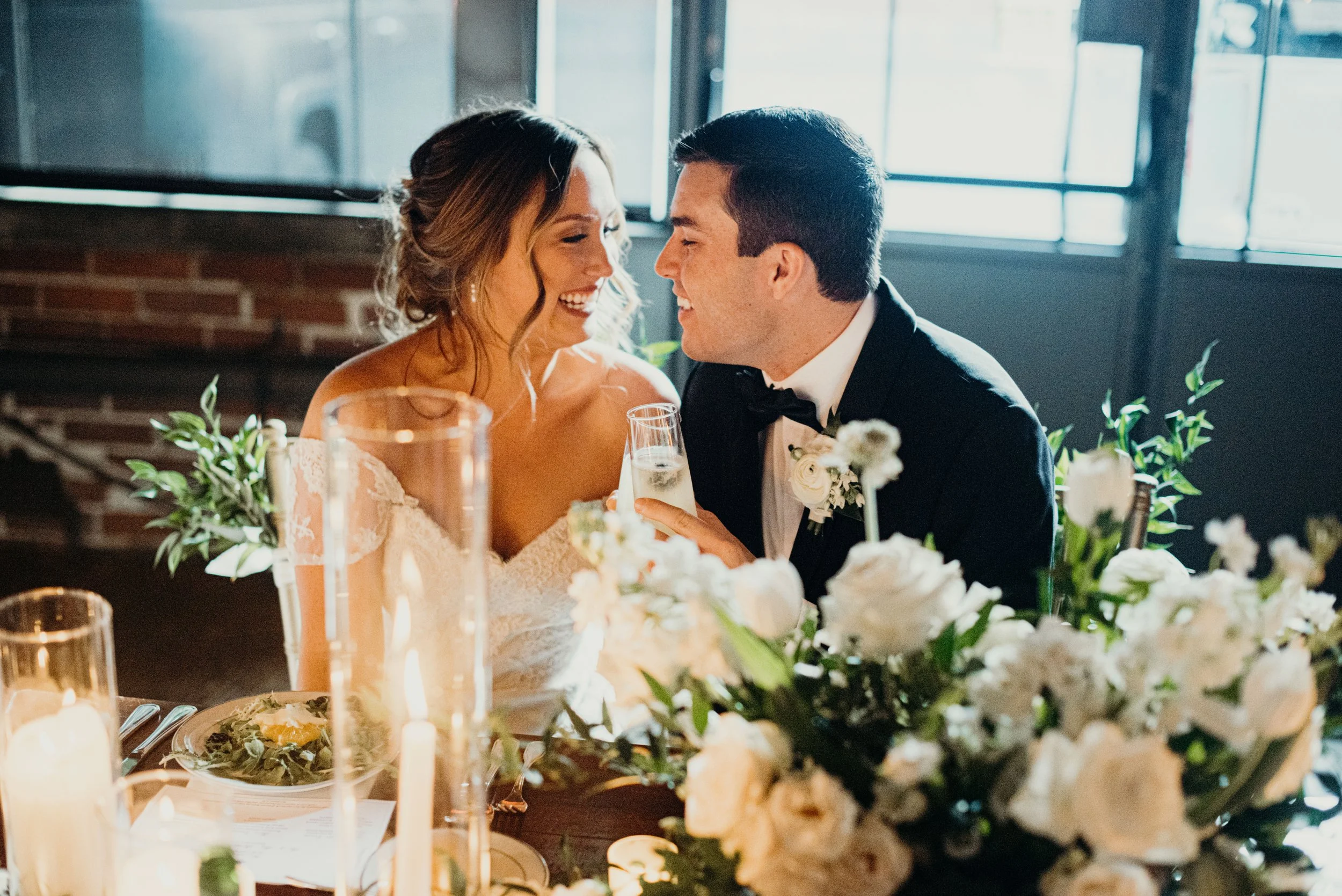 A bride and groom sitting closely at a decorated wedding reception table with flowers and candles, smiling and gazing at each other.