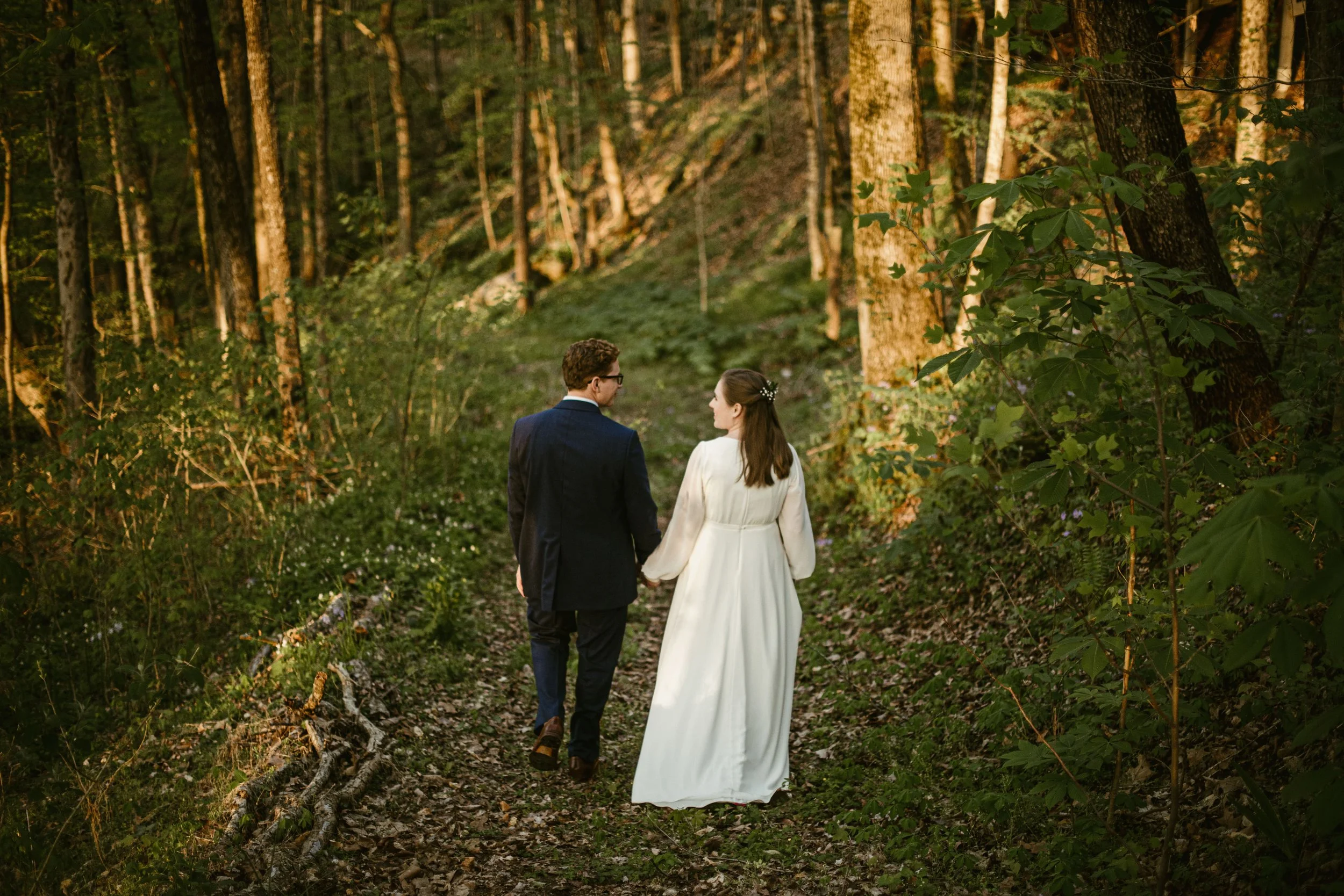 A bride and groom holding hands and walking through a forested area with tall trees and green foliage.