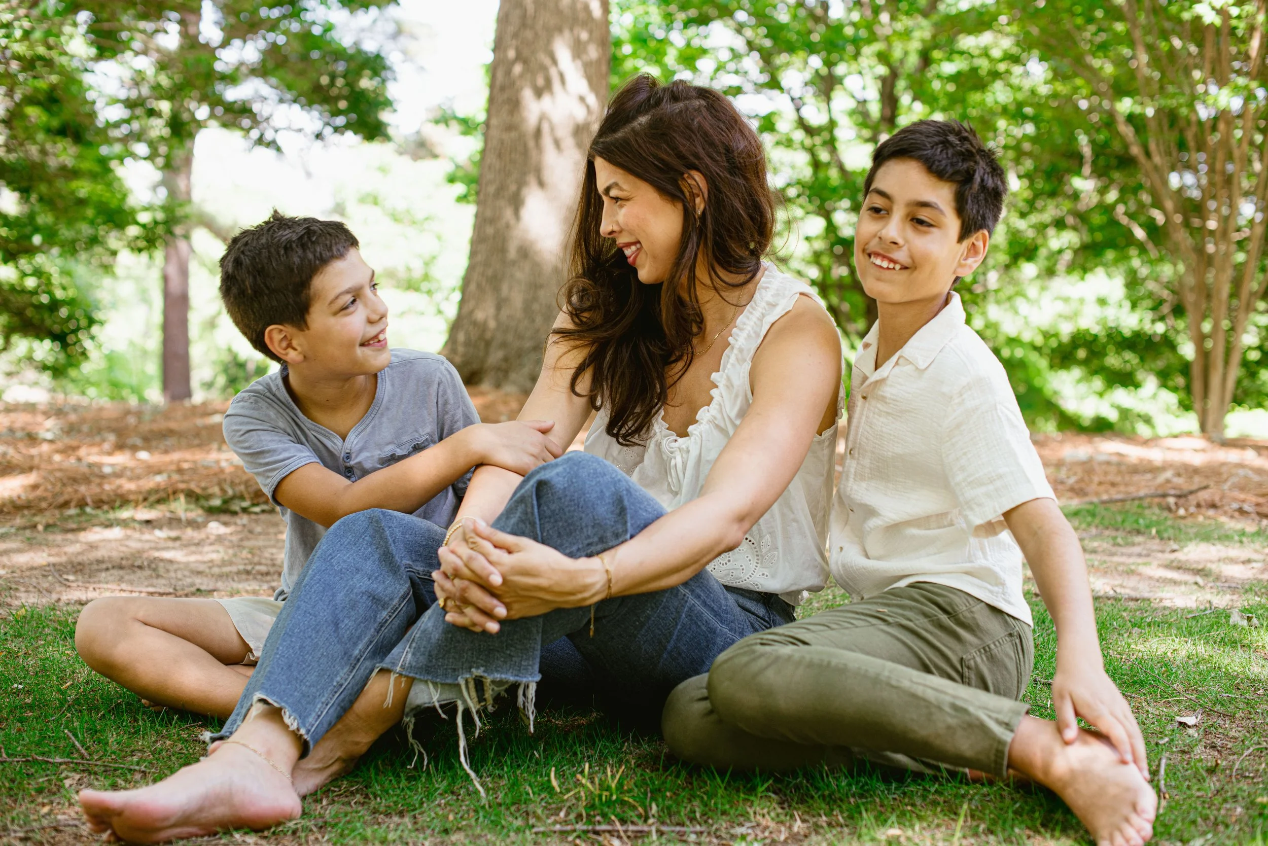 A woman and two boys sitting outdoors on grass, smiling and engaging with each other in a wooded park setting.