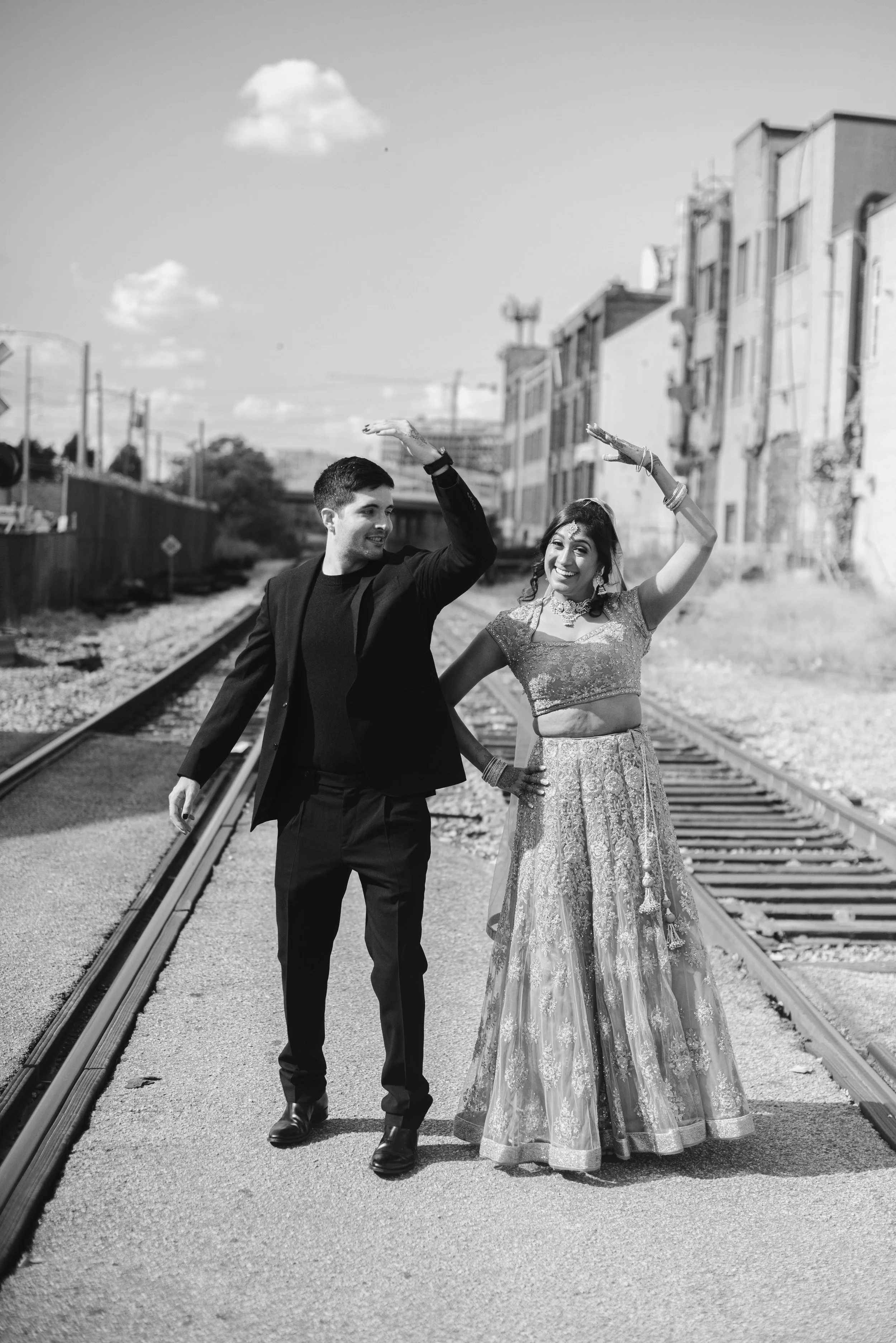 A happy couple dressed in traditional Indian wedding attire walking along train tracks outdoors, smiling and raising their arms in celebration.