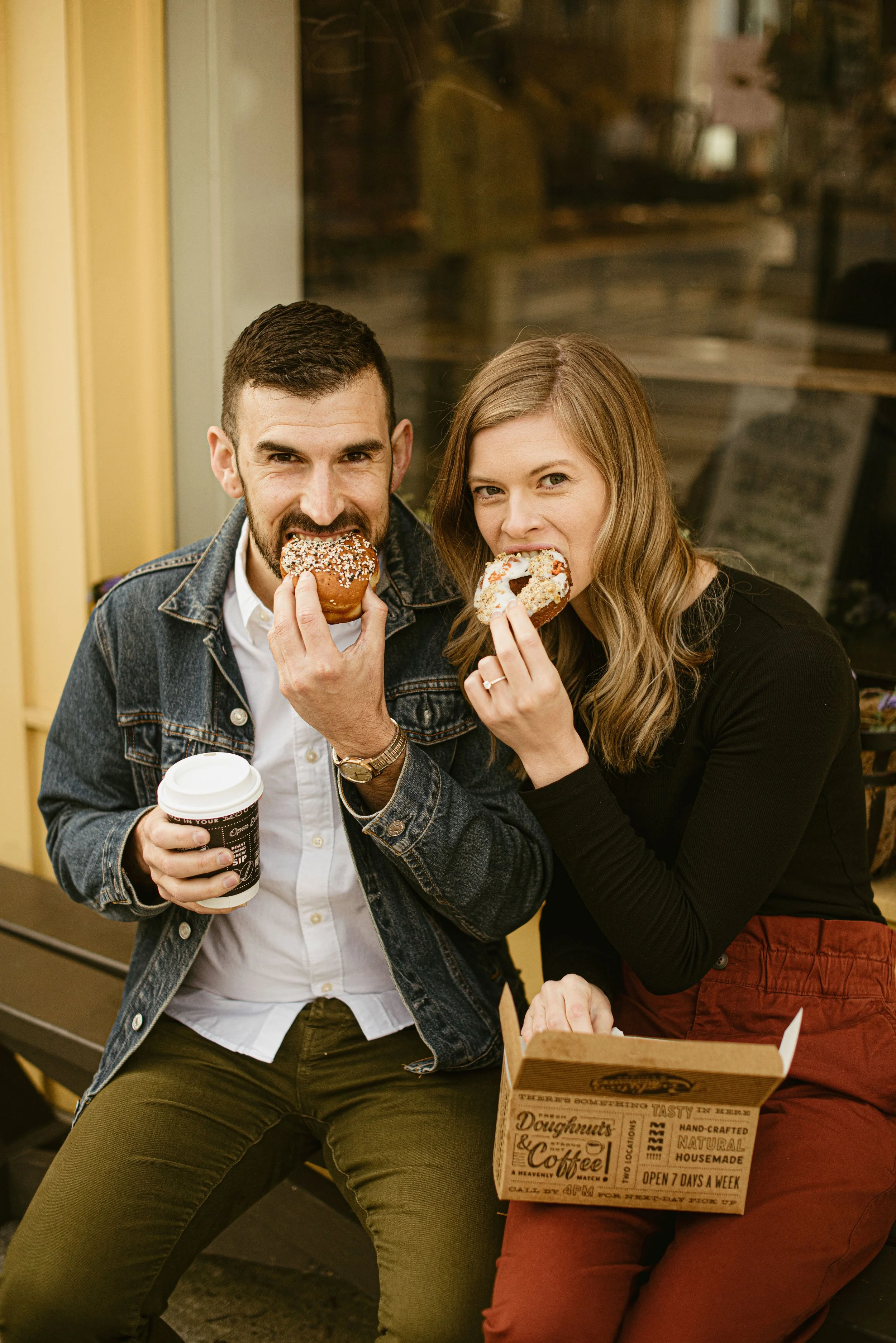 A man and woman are sitting together, eating donuts. The man has dark hair and a beard, and is wearing a denim jacket and holding a coffee cup. The woman has long, light brown hair and is wearing a black top, with a ring on her finger, and is holding