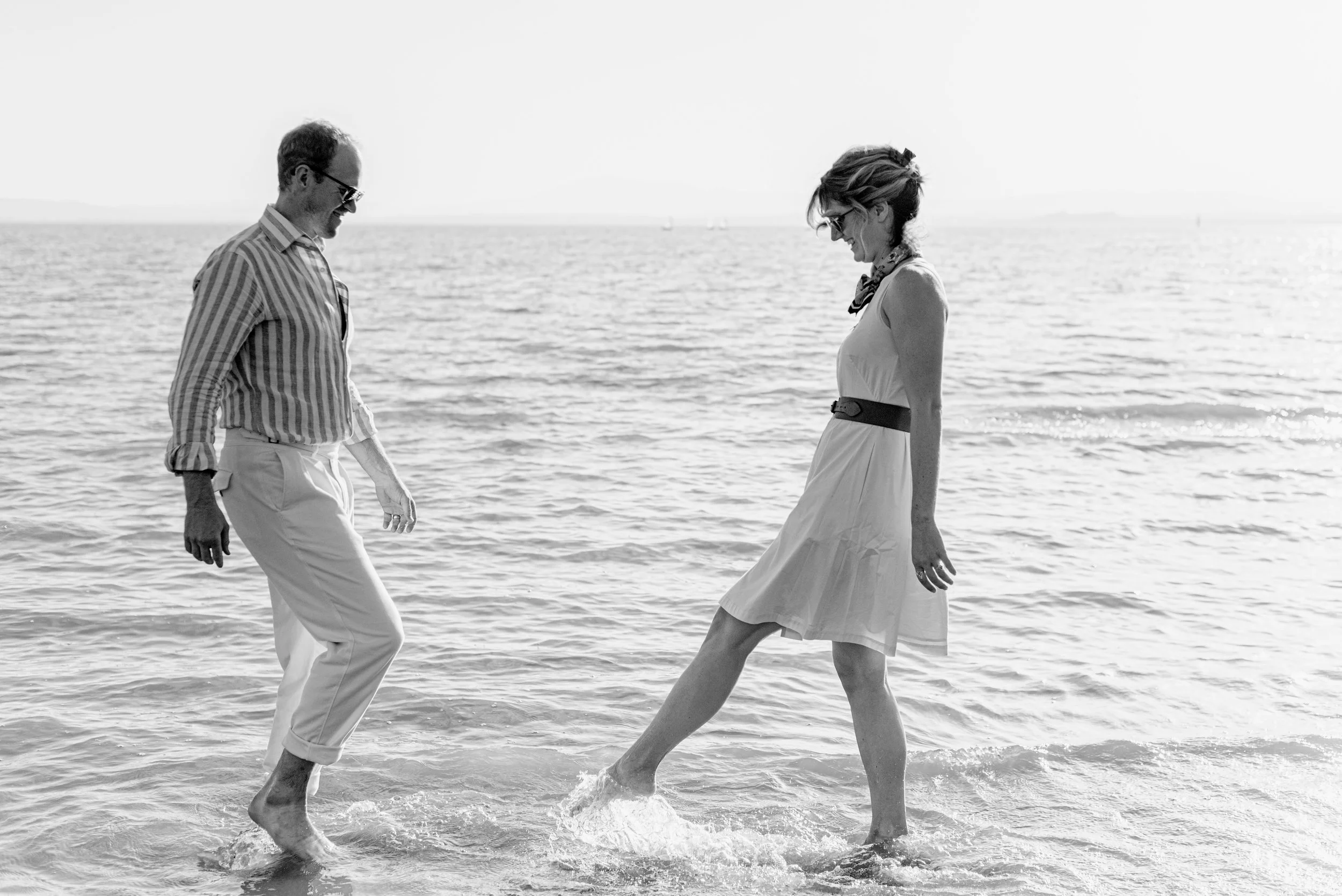 A black and white photo of a man and a woman smiling and walking in the water at the beach. The man is wearing striped shirt and glasses, and the woman is in a sleeveless dress with a scarf, and they are looking at each other.