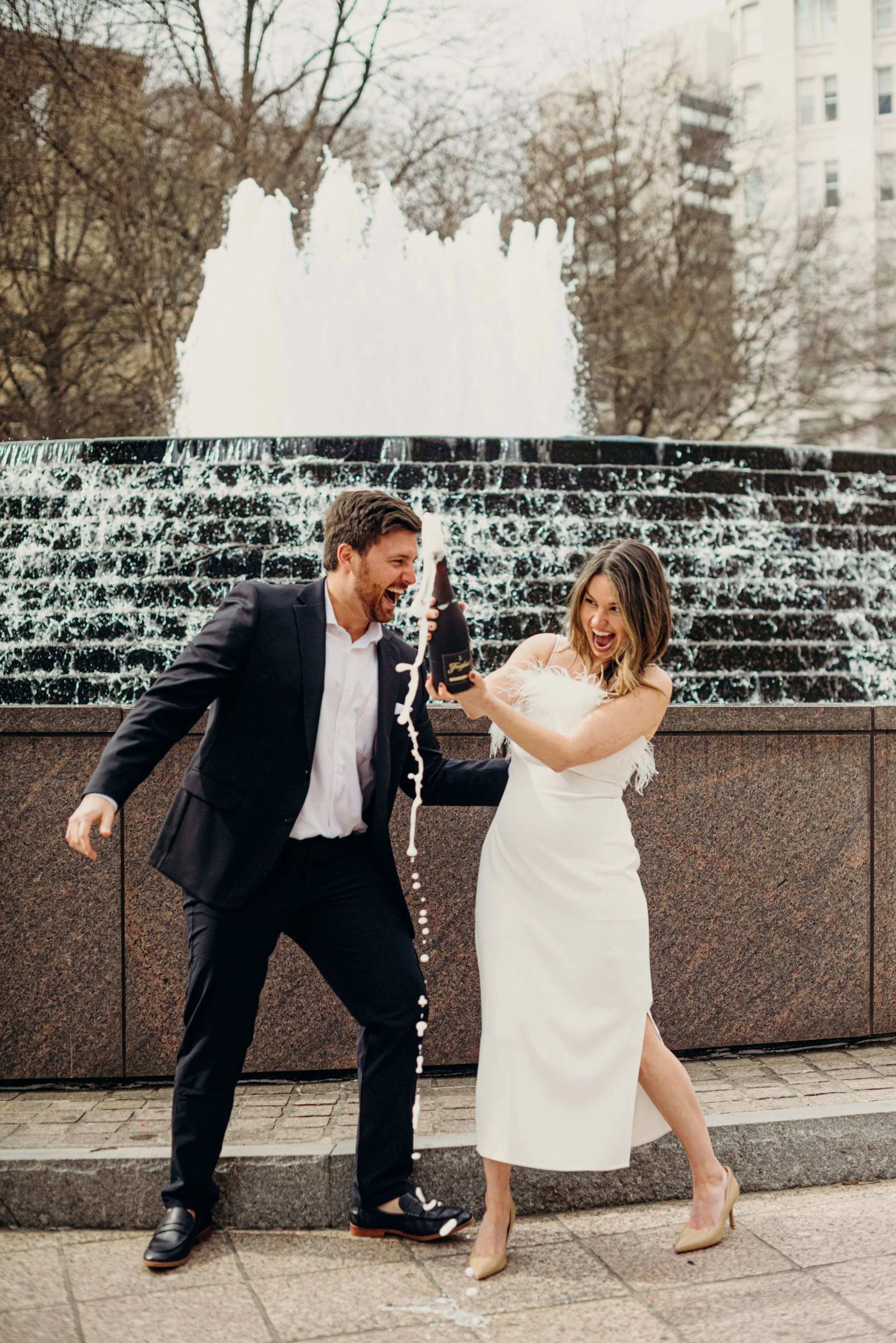 Man and woman celebrating with champagne by a fountain in an urban park, woman in a white dress and heels, man in a black suit, both smiling and laughing, confetti falling.