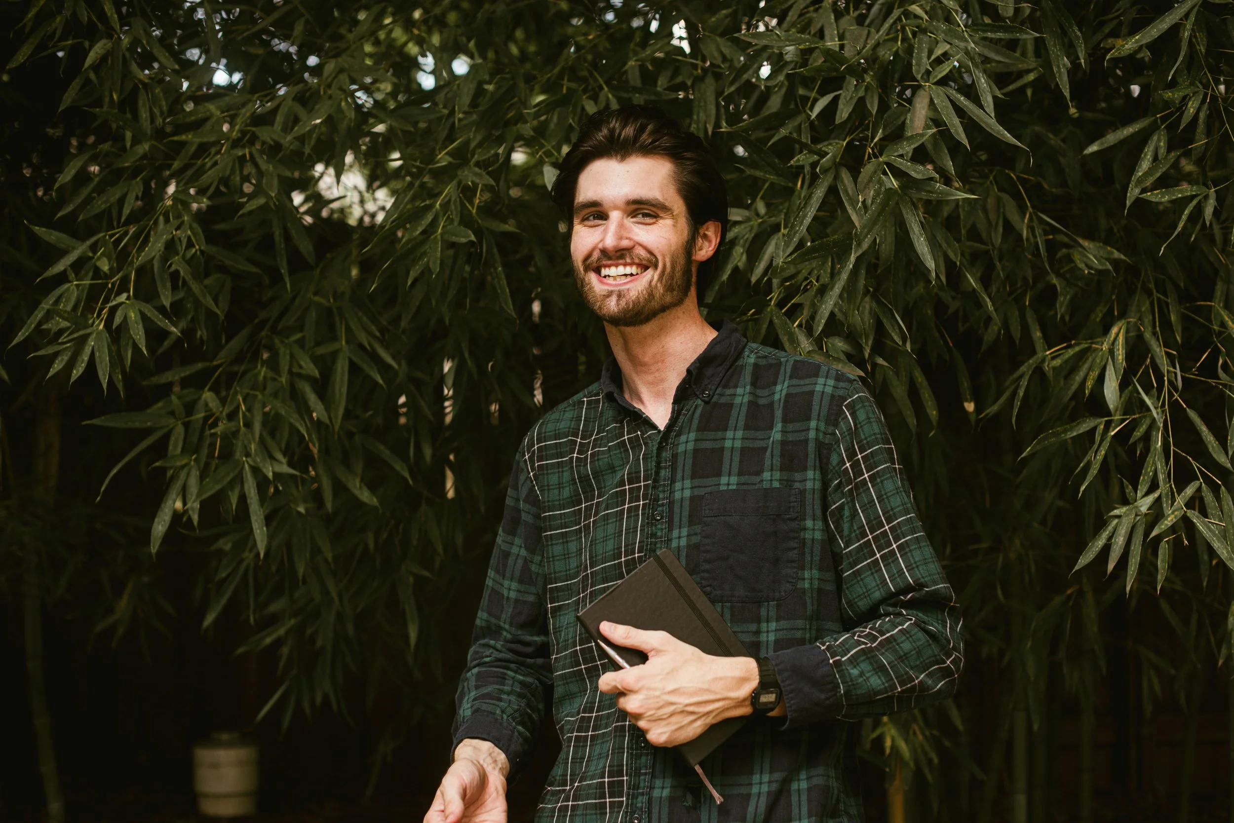 A smiling man with dark hair and a beard, wearing a green and black plaid shirt, holding a tablet or notebook, standing in front of lush green foliage.