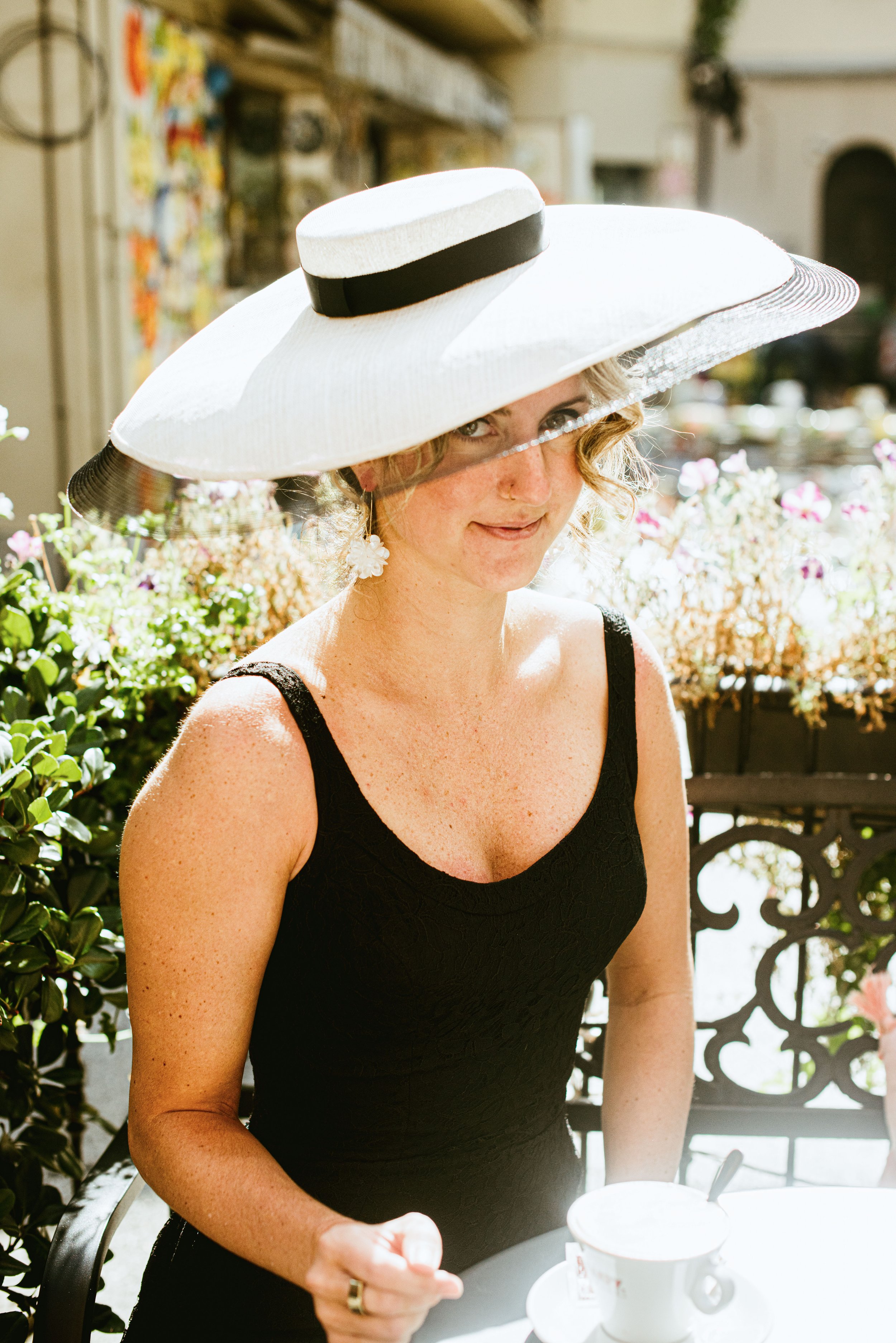 A woman wearing a wide-brimmed white hat with a black band, sitting outdoors at a table with flowers in the background, holding a spoon and a cup of coffee.