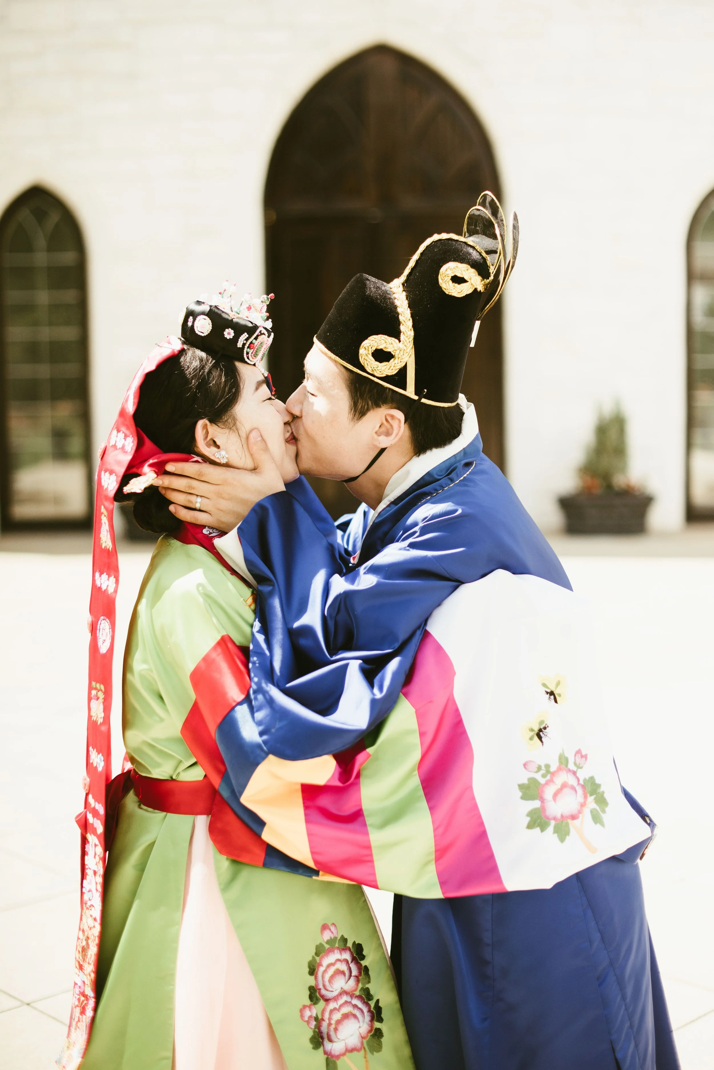 A couple in traditional Korean hanbok dress sharing a kiss, with the woman wearing a tiara and the man wearing a black and gold traditional hat, standing outside in front of a white building with arched windows.