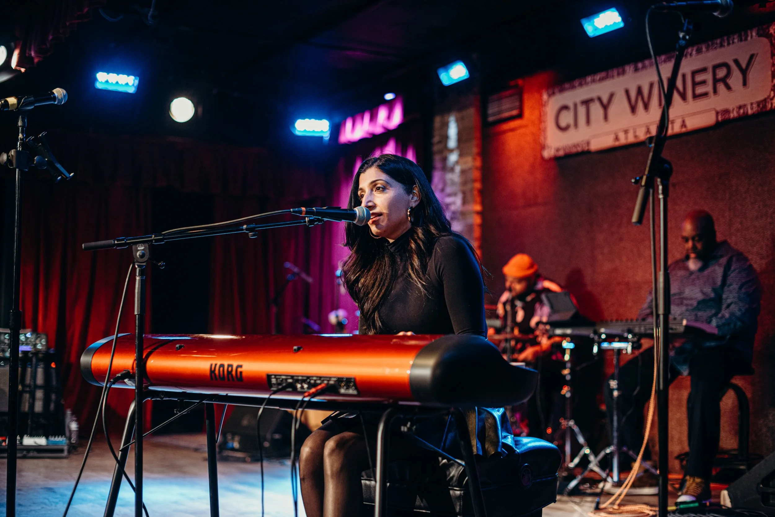 A woman playing keyboard on stage with microphones and musical equipment, with a sign reading 'City Winery Atlanta' in the background.