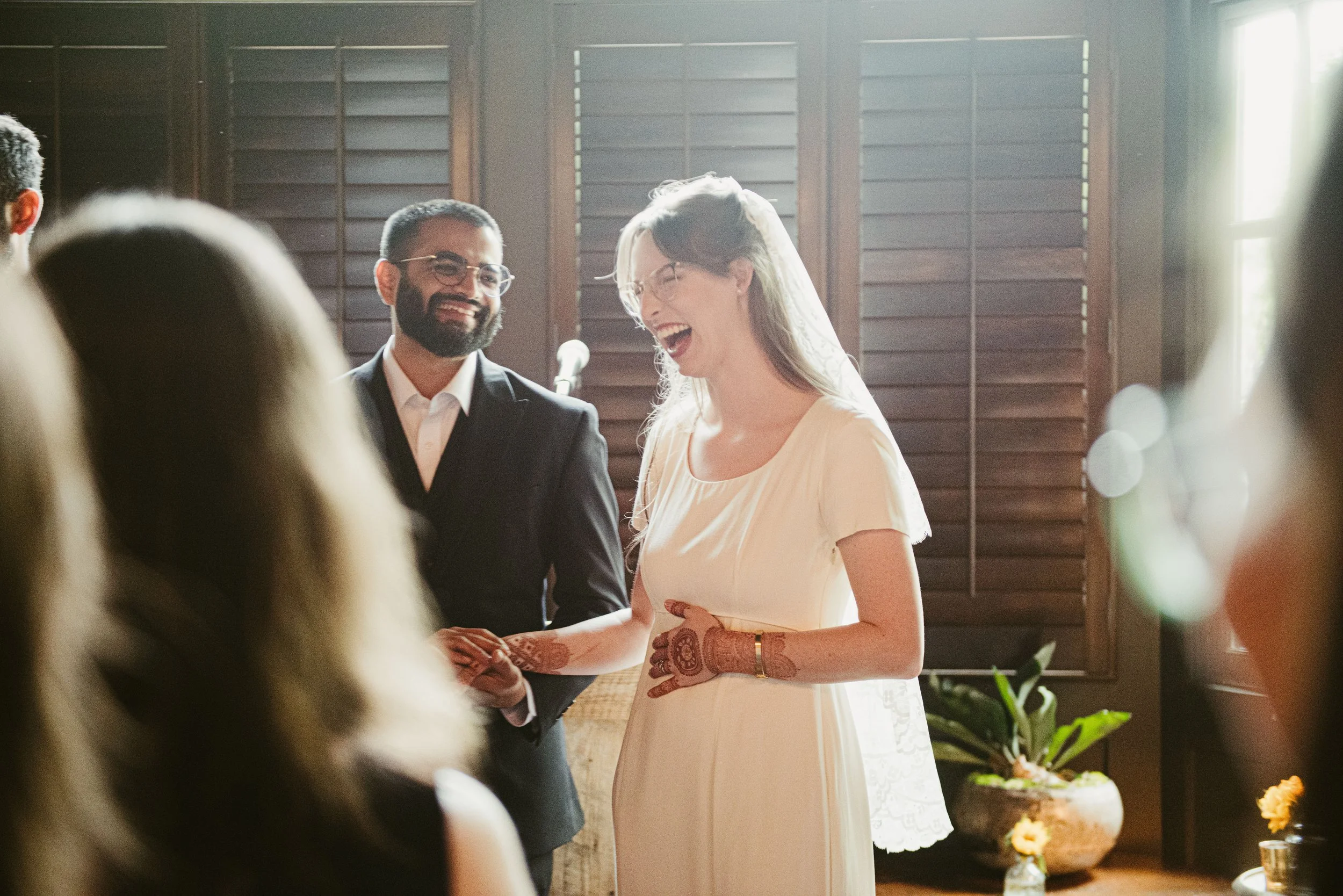 A bride and groom are exchanging vows at their wedding ceremony, smiling and holding hands, with guests visible in the foreground.