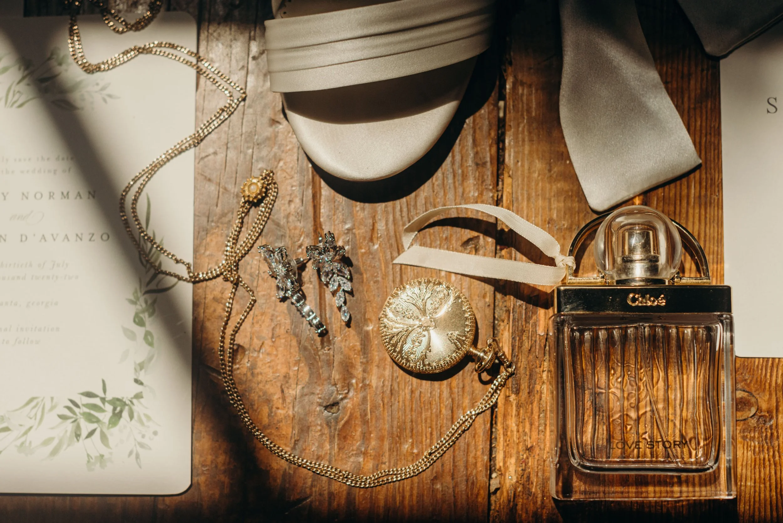 A flat lay of jewelry, perfume, and accessories on a wooden surface, including pearl necklaces, earrings, a gold pendant, a beige bow-tied box, and a perfume bottle labeled 'Chloé'.
