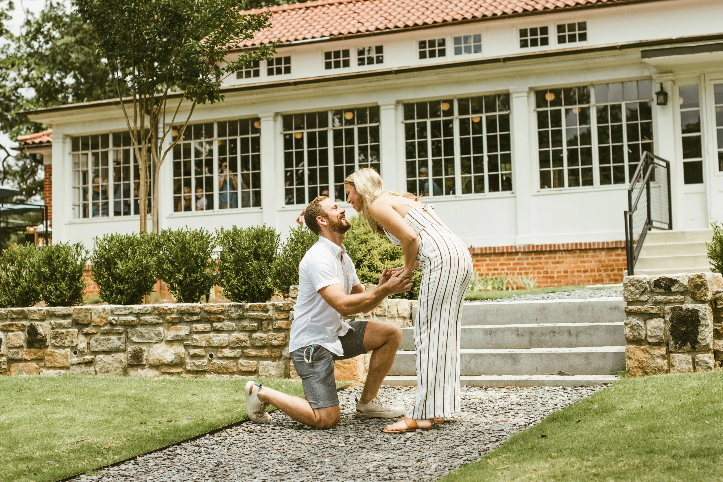 A man kneeling on one knee proposing to a woman on a grassy outdoor area in front of a white house with large windows. The woman is smiling and wearing a striped dress, while the man wears a white shirt and shorts. Two people are visible behind the w