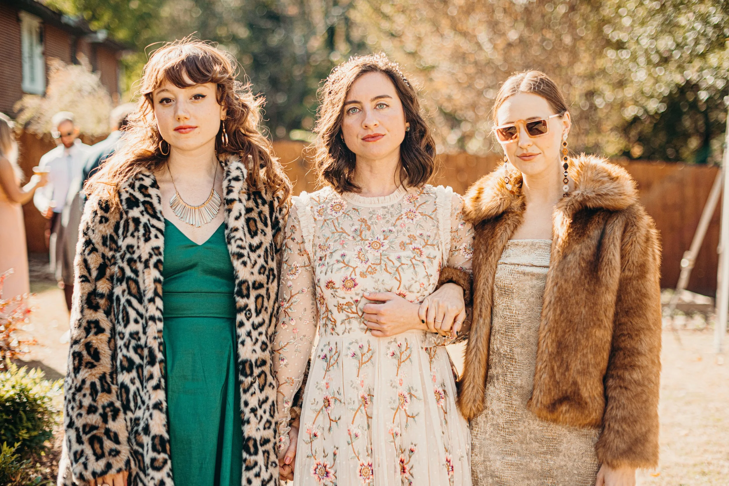 Three women standing outdoors in sunlight, dressed in stylish clothes and accessories, with two more people blurred in the background.