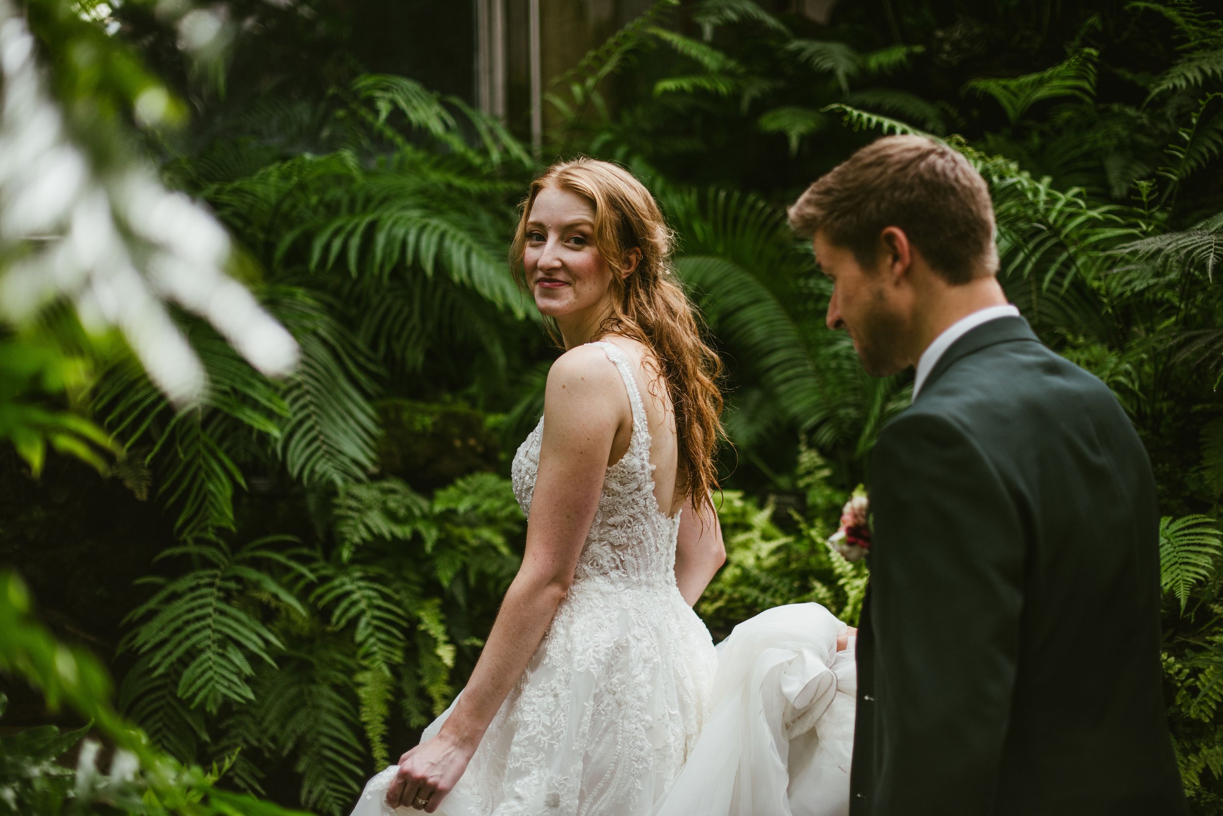 A bride in a white lace wedding dress and a groom in a dark suit standing in a lush green garden.