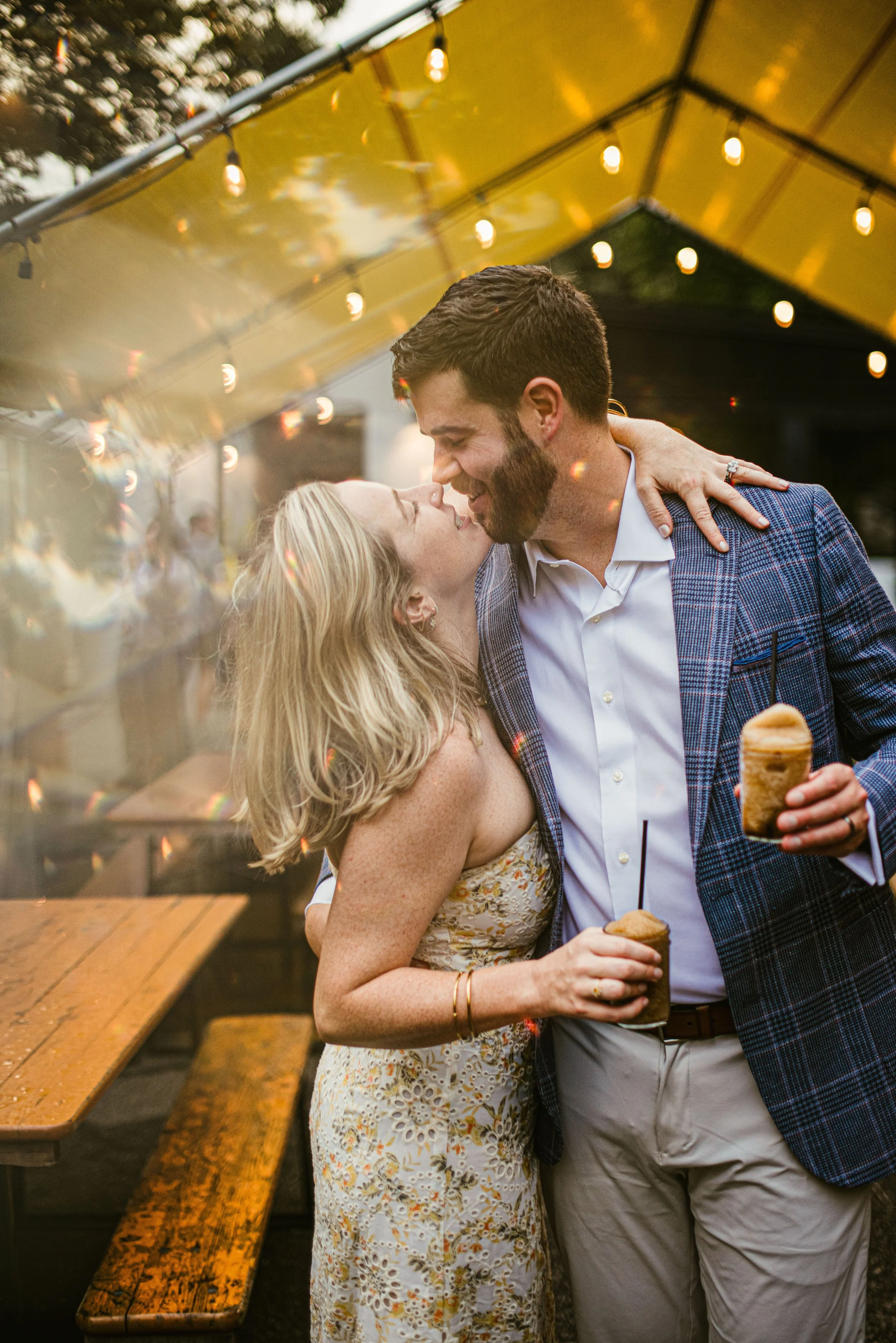 A smiling couple is sharing a tender moment, about to kiss, at an outdoor event during the evening, with string lights overhead and holding ice cream cones.