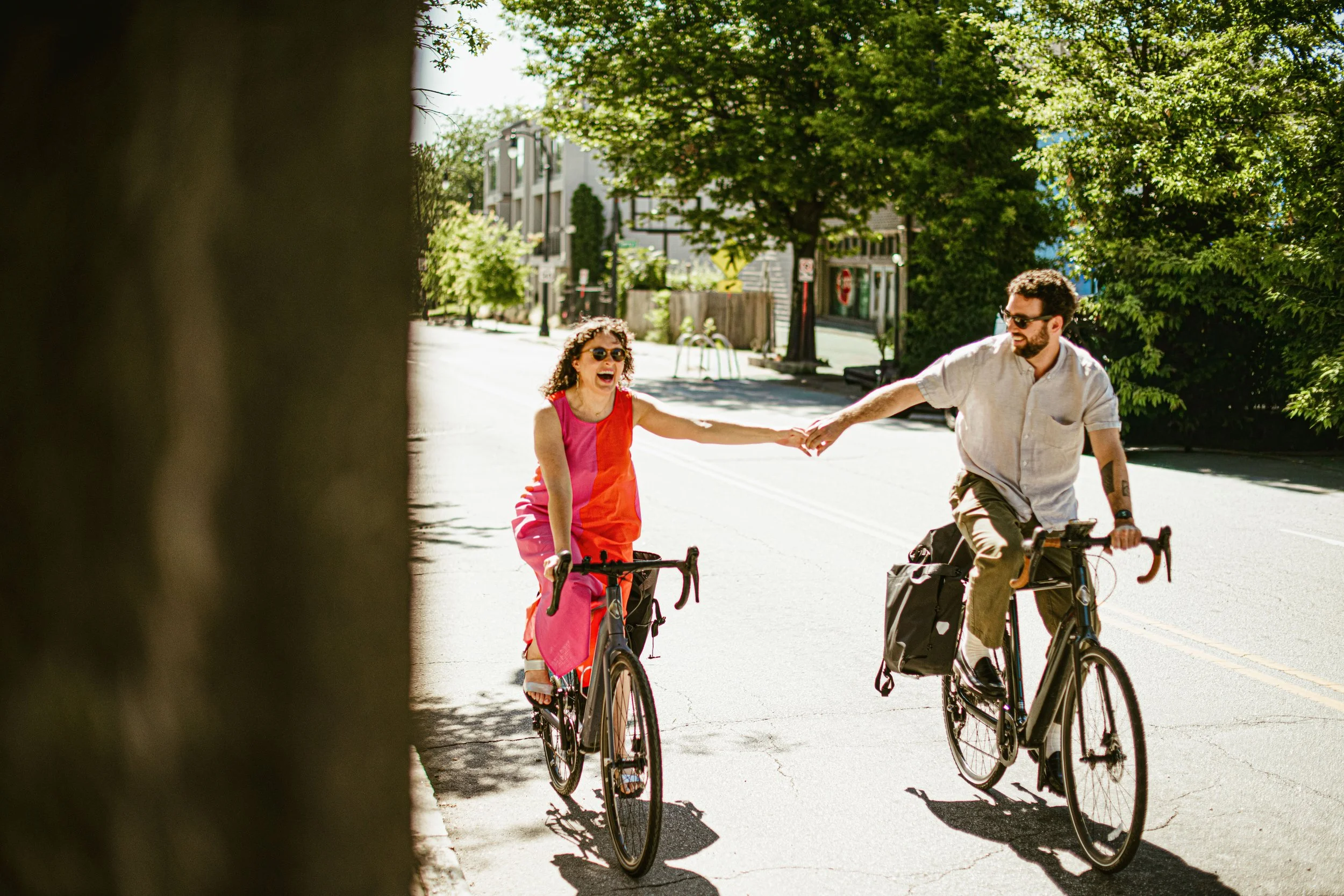 Two people biking on a sunny street, laughing and holding hands, with green trees and buildings in the background.