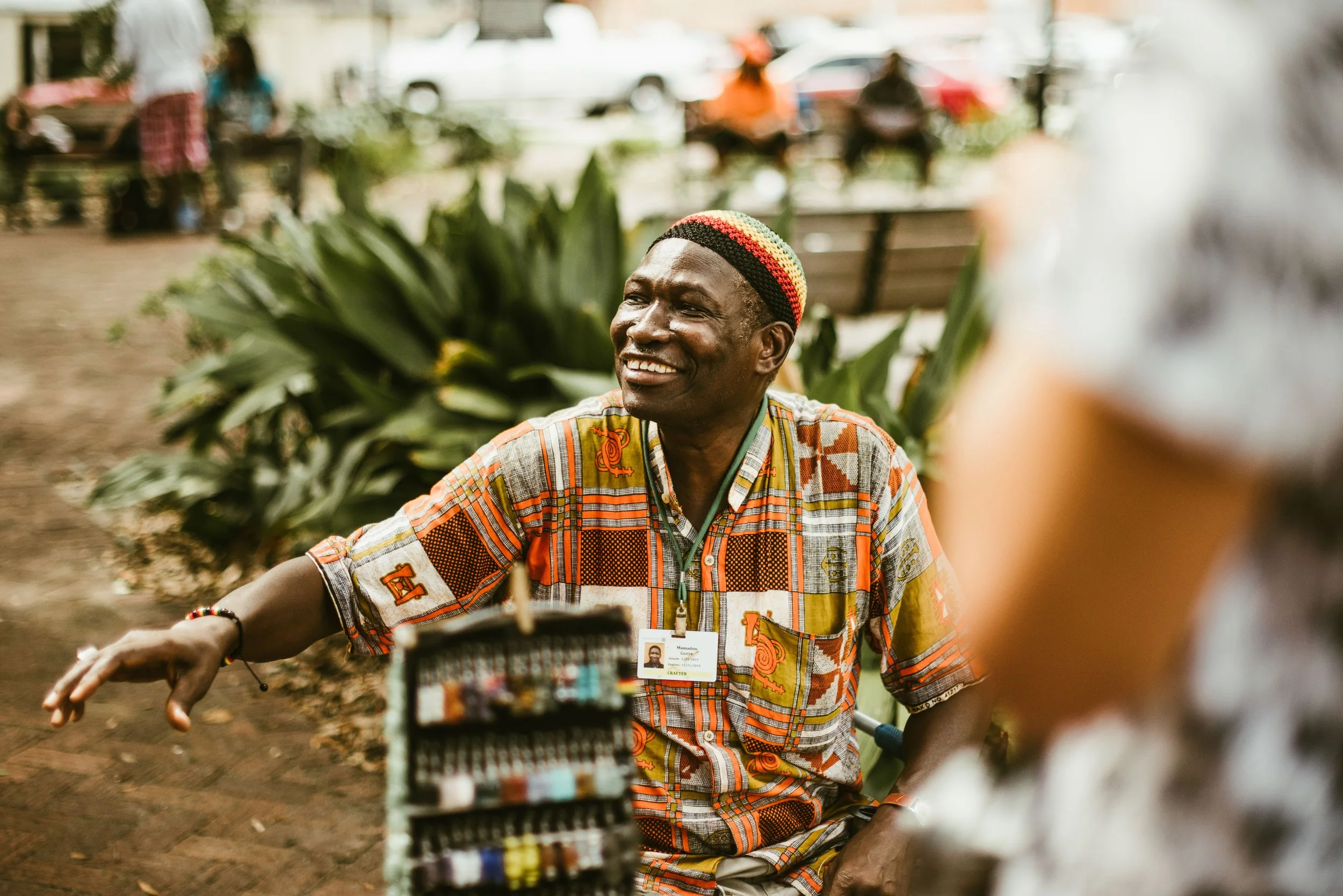 A smiling man in colorful, patterned clothing and a knit hat with red, yellow, and green stripes, sitting outdoors with plants and blurred people and cars in the background.