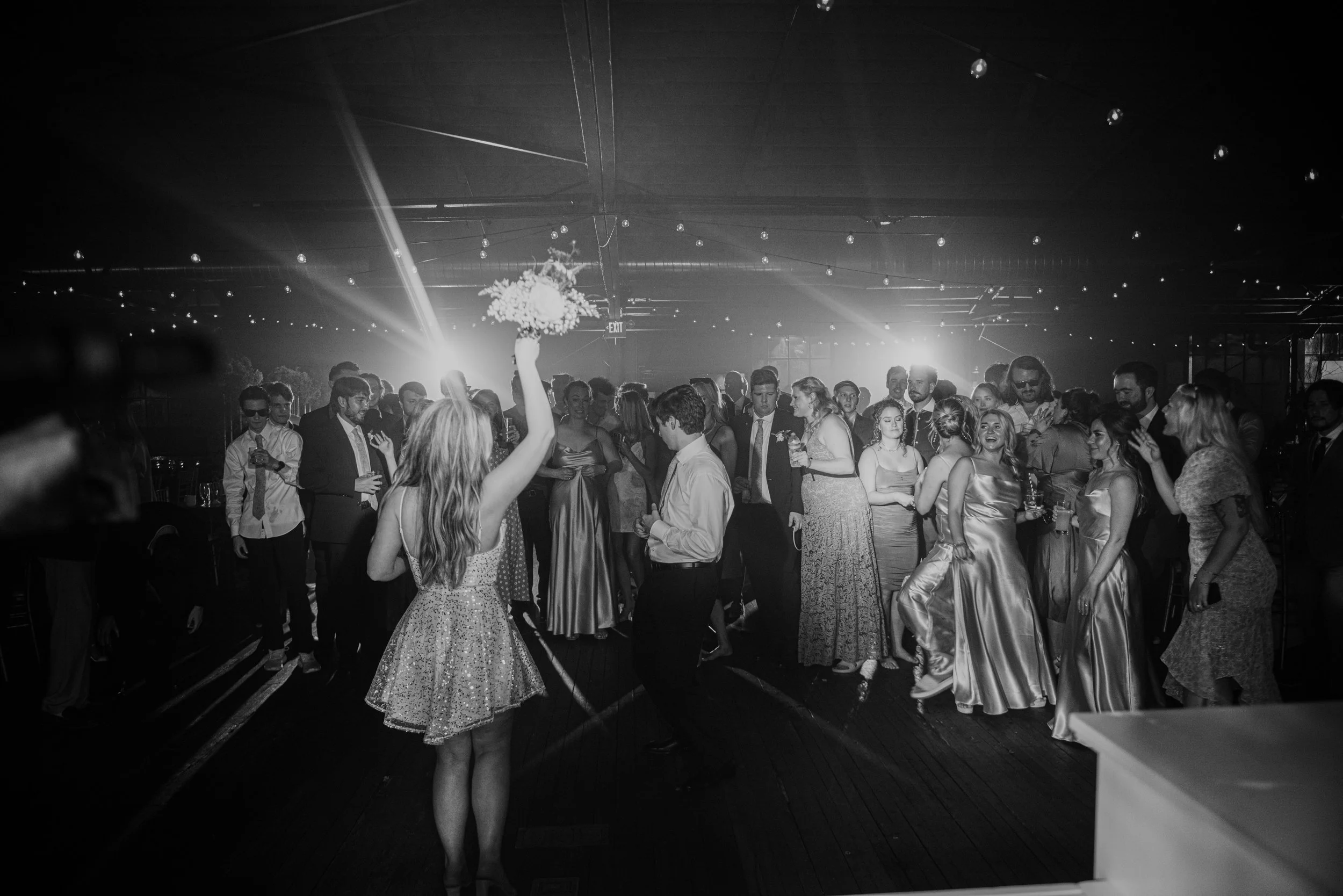People at a party, with a woman holding a bouquet of flowers raised in the air, dancing, and others socializing and enjoying drinks. The setting appears to be an indoor event space with dim lighting and string lights on the ceiling.