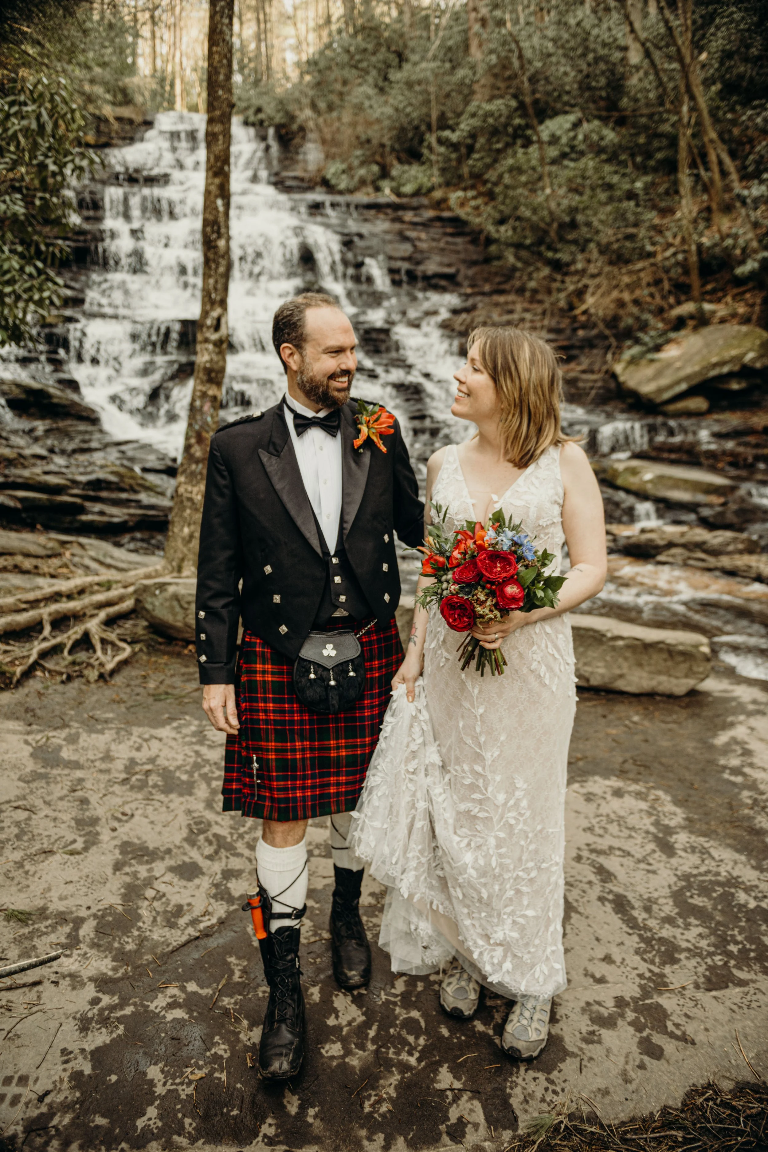 A couple in wedding attire standing outdoors in front of a waterfall in a forest. The groom is dressed in a traditional Scottish outfit with a kilt, jacket, and bow tie, while the bride is wearing a lace wedding dress and holding a bouquet of red and
