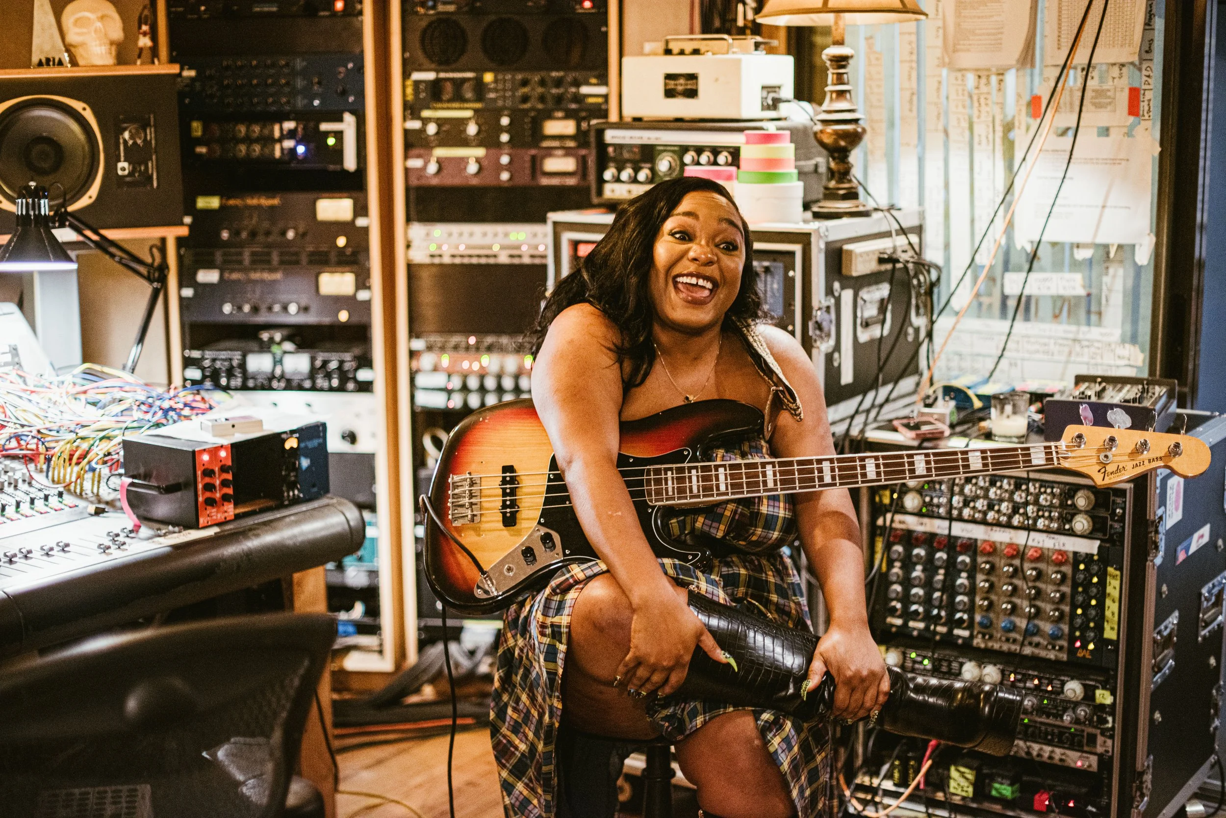 A woman with dark hair and a plaid skirt playing an electric guitar in a music studio filled with audio equipment, wires, and colorful tape rolls.