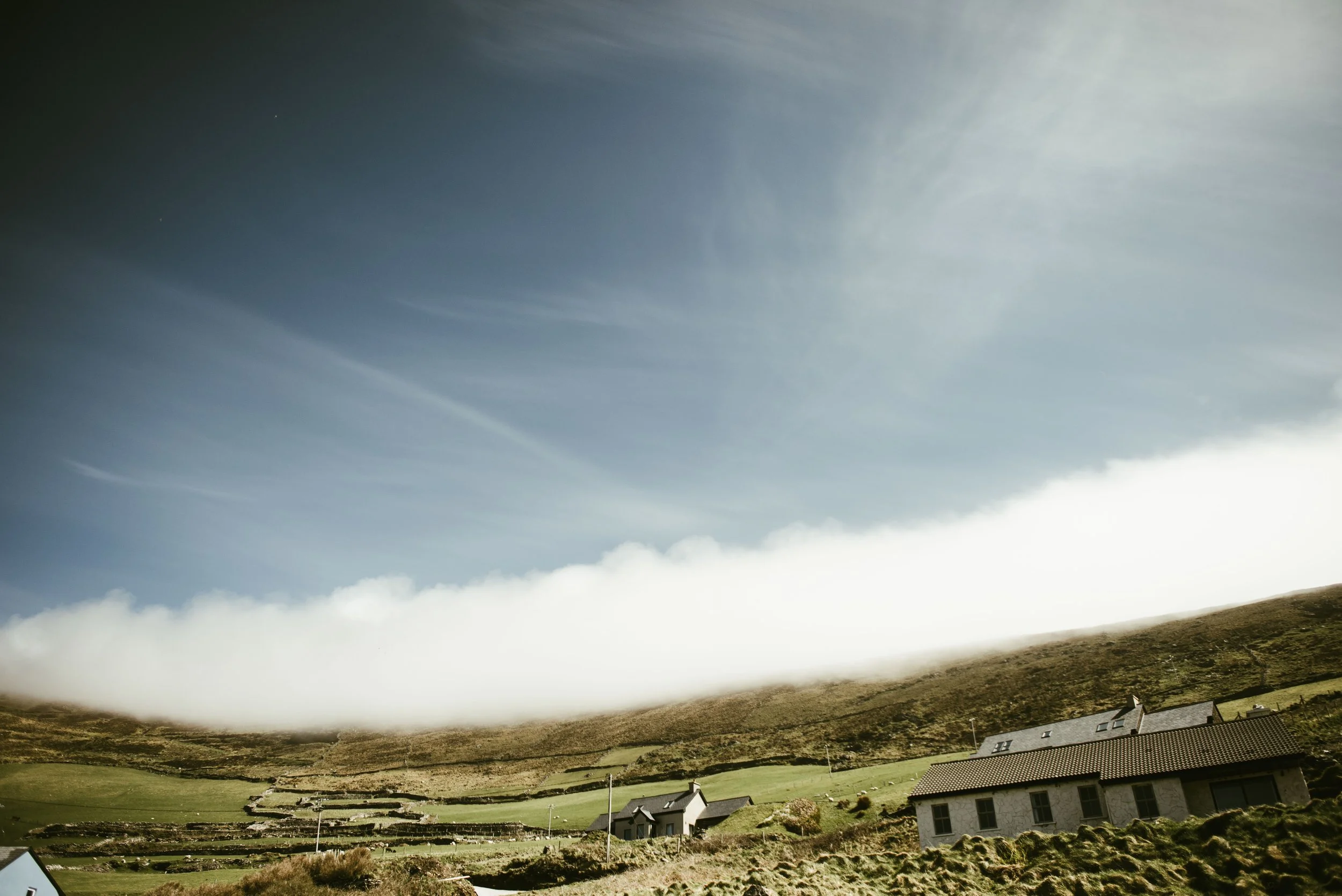 A landscape with green grassy hills, houses with gray roofs, and a sky with clouds and sunlight in Ireland
