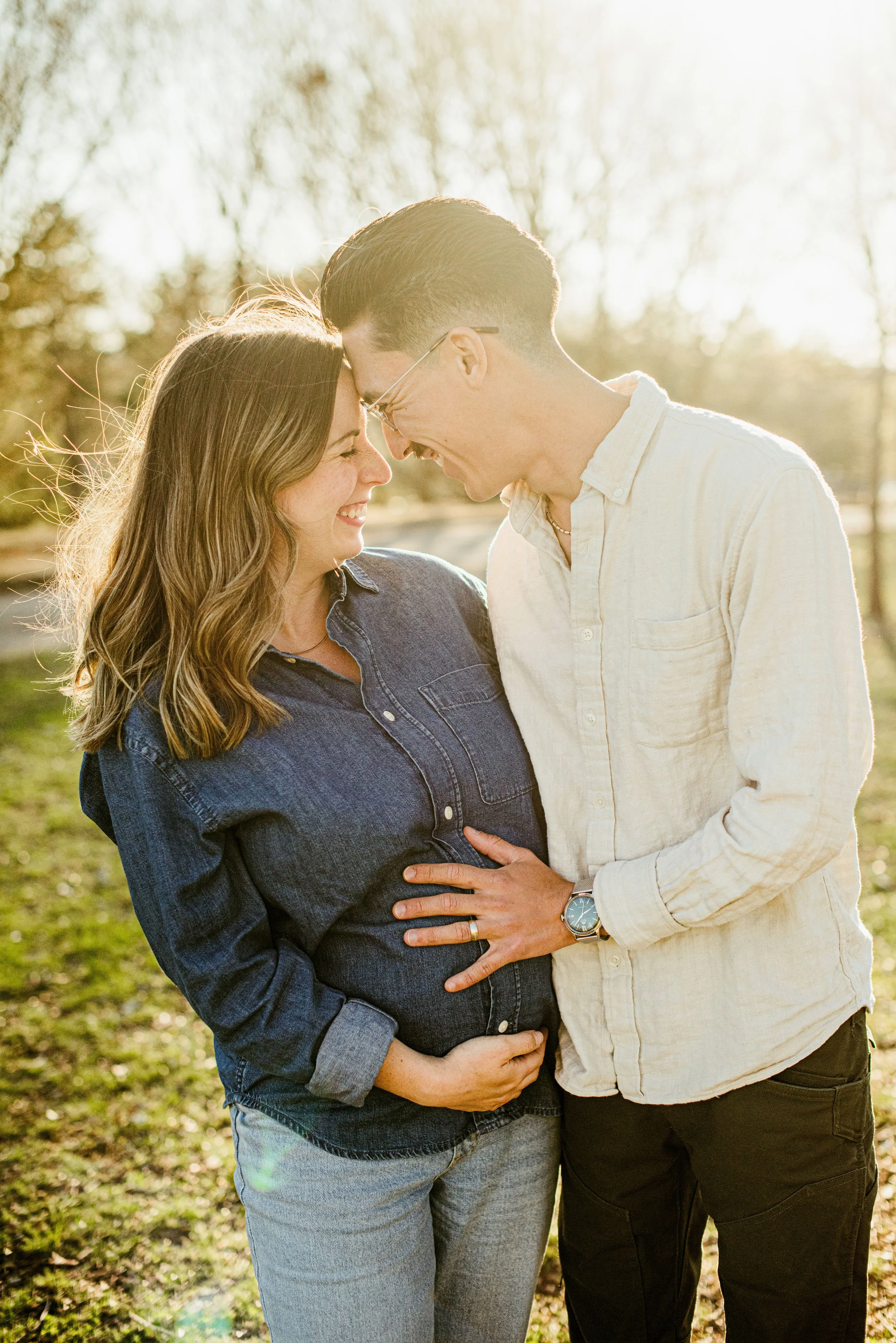 A couple is standing outdoors, touching foreheads and smiling, with sunlight shining behind them. The woman is pregnant and wearing a denim shirt, while the man is dressed in a light-colored shirt and glasses.