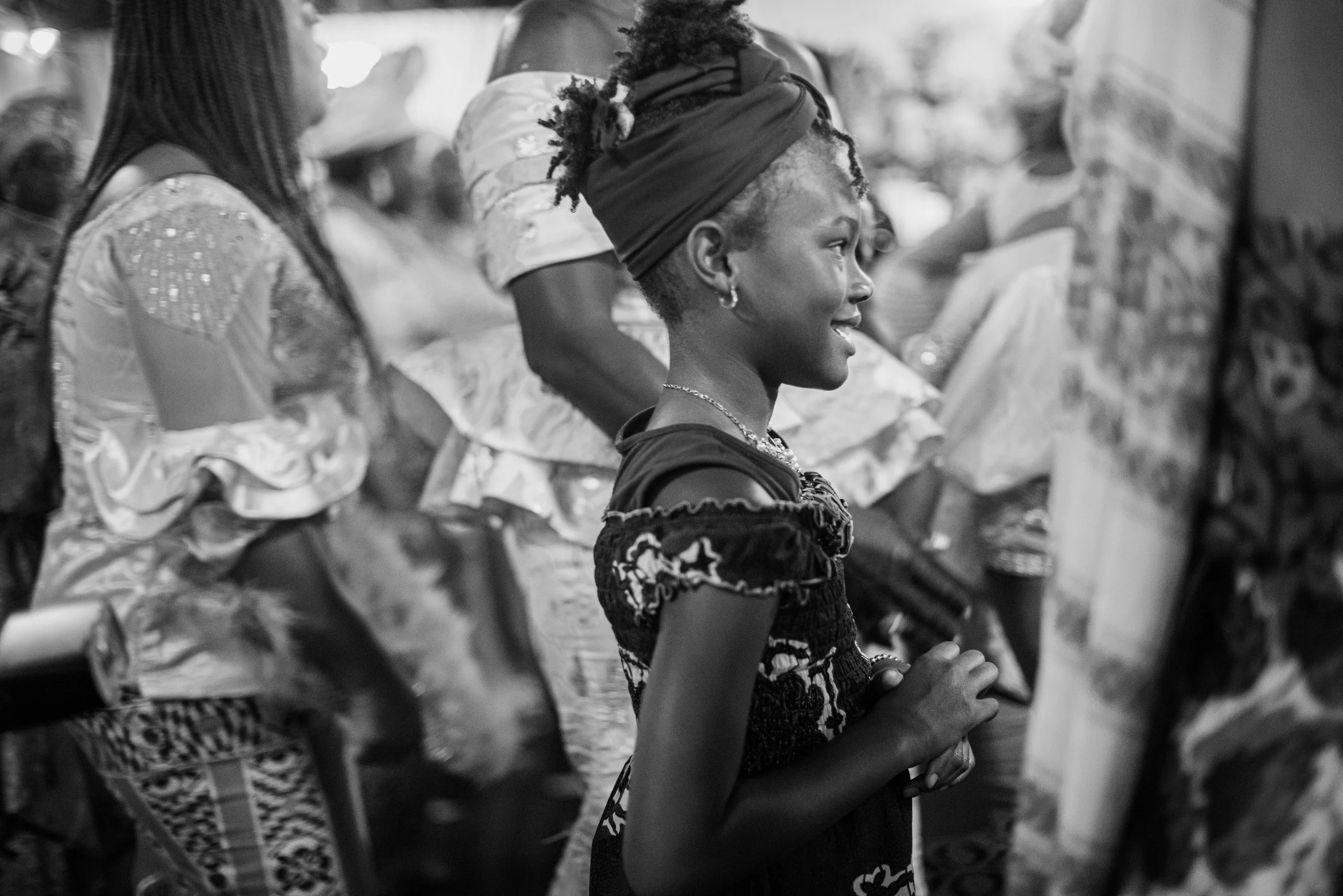 A group of women standing in line, with one woman in the center wearing a headwrap and patterned dress, clasping her hands together in prayer or contemplation.