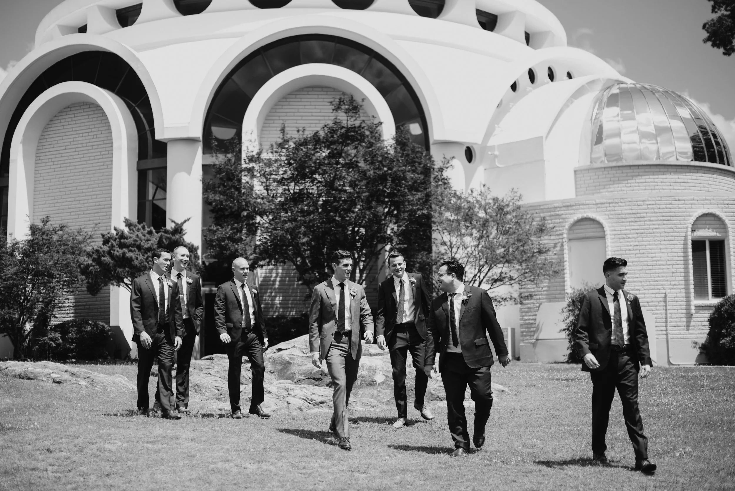 A group of men in suits walking outdoors in front of a modern building with large round windows and a dome structure, with trees and rocks in the background.
