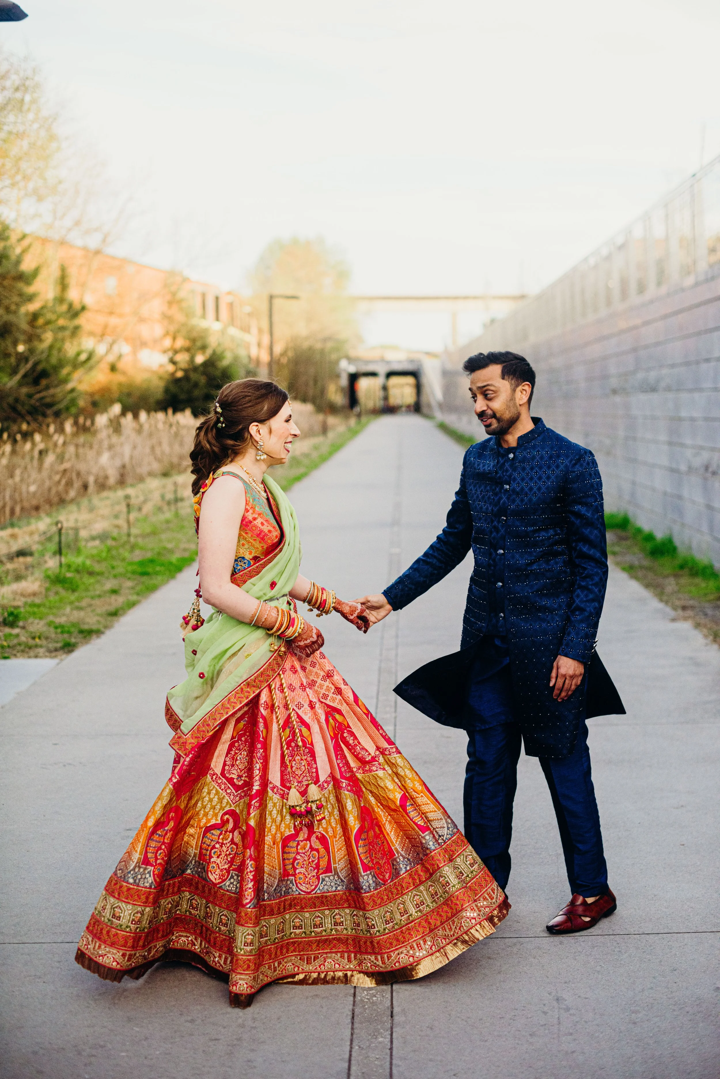 A couple dressed in colorful traditional Indian attire holding hands on a city sidewalk with trees and buildings in the background.