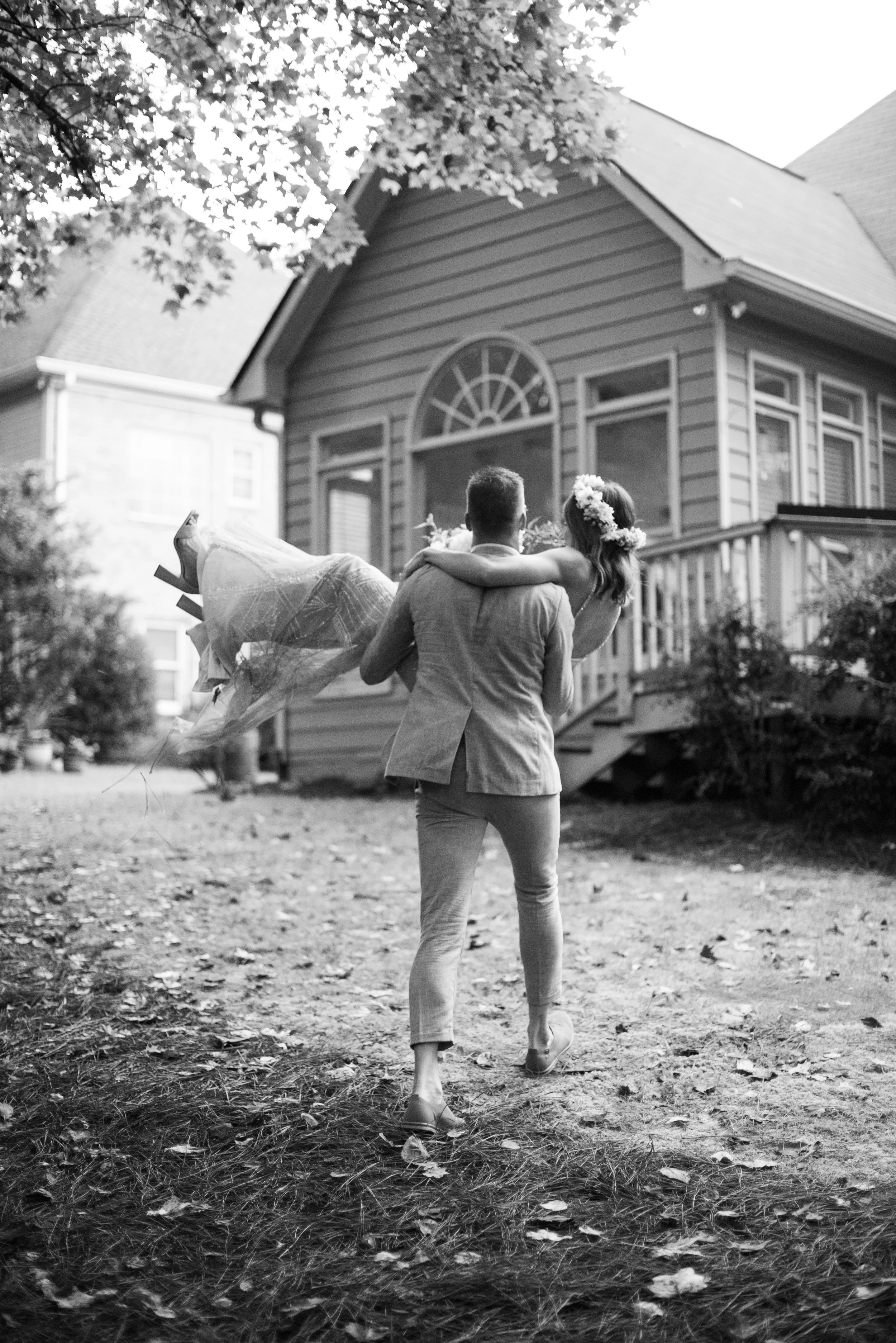A couple dressed in formal attire, with the woman wearing a flower crown, sharing a dance or embrace outdoors near a house, in a black-and-white photo.