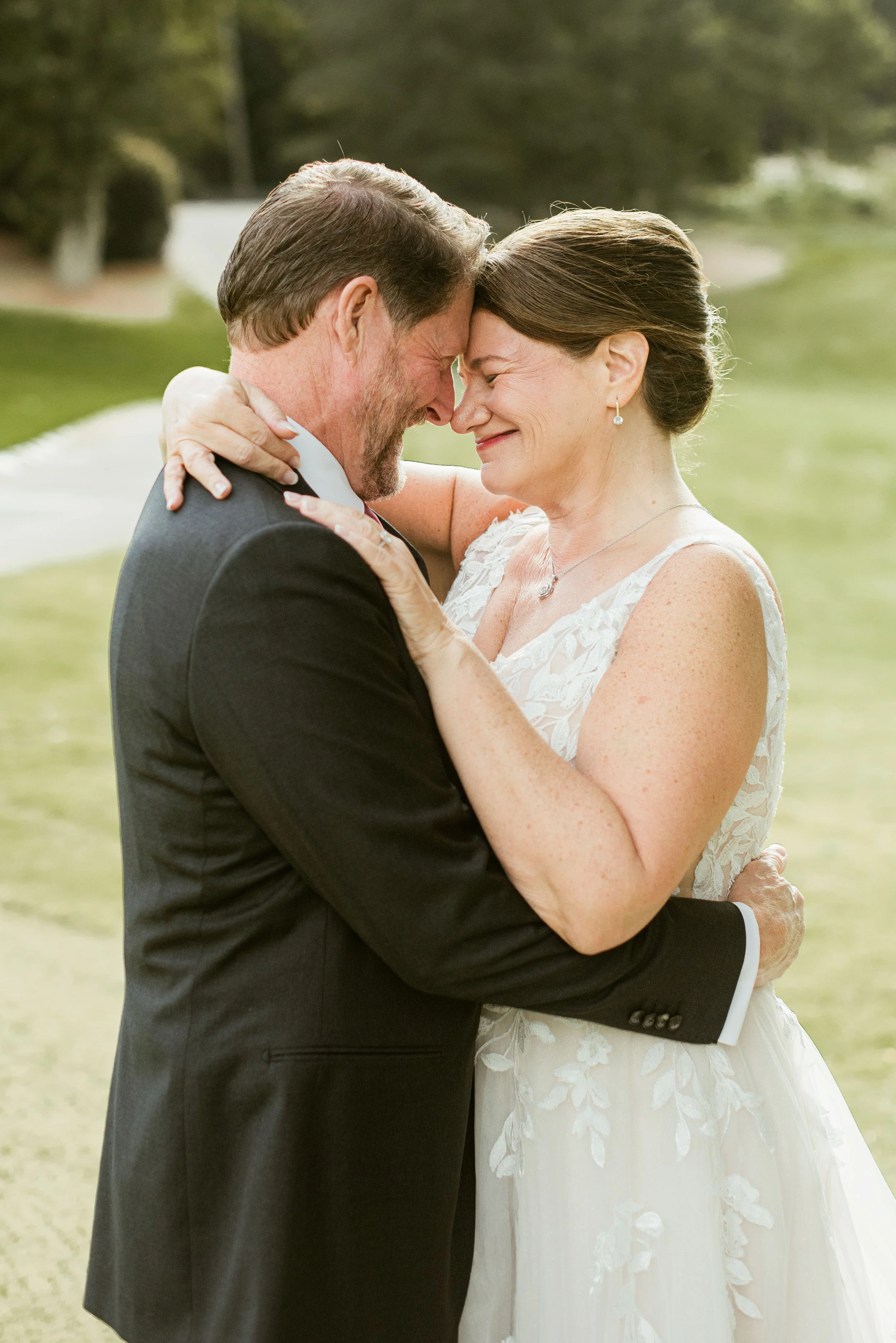 A loving couple, a man in a dark suit and a woman in a white lace wedding dress, holding each other with foreheads touching outdoors near a body of water and greenery.