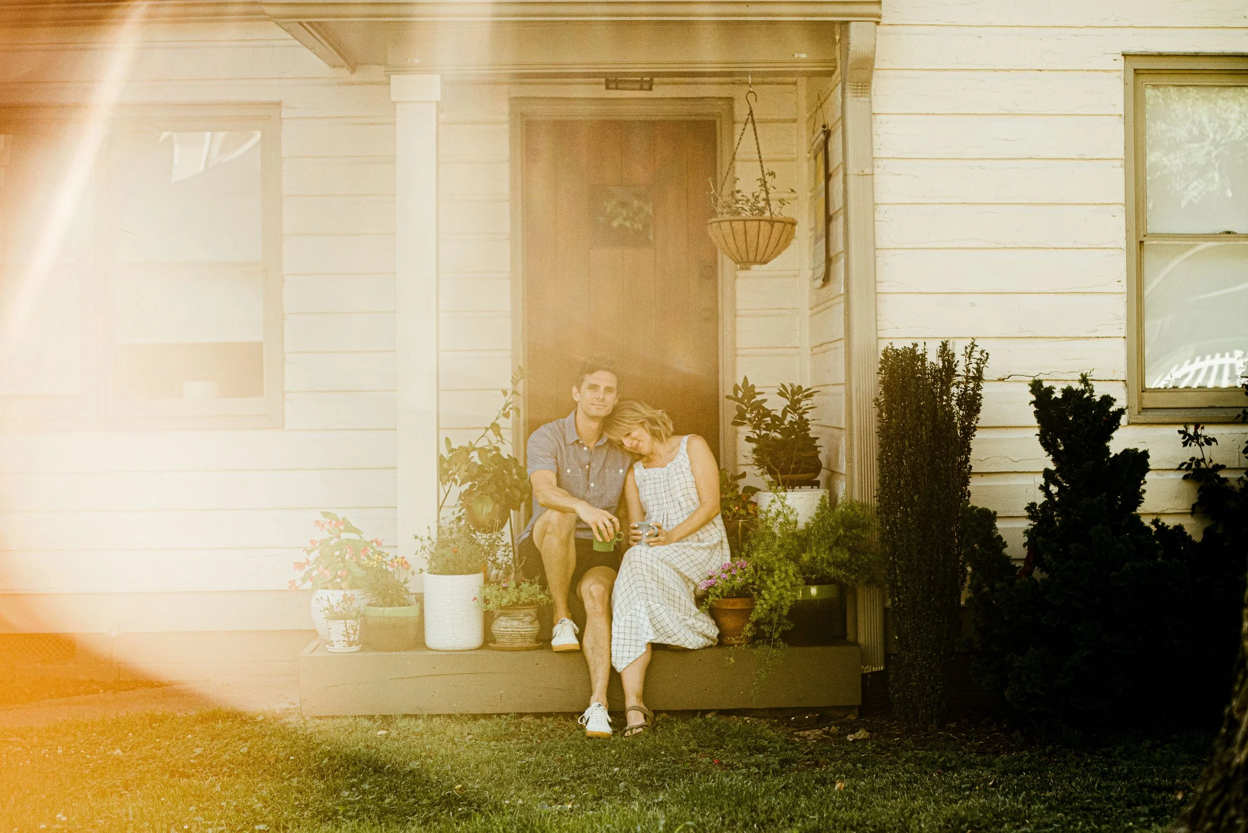 A young couple sitting on a porch outside a white wooden house, surrounded by potted plants and bushes, with sunlight creating a warm glow.