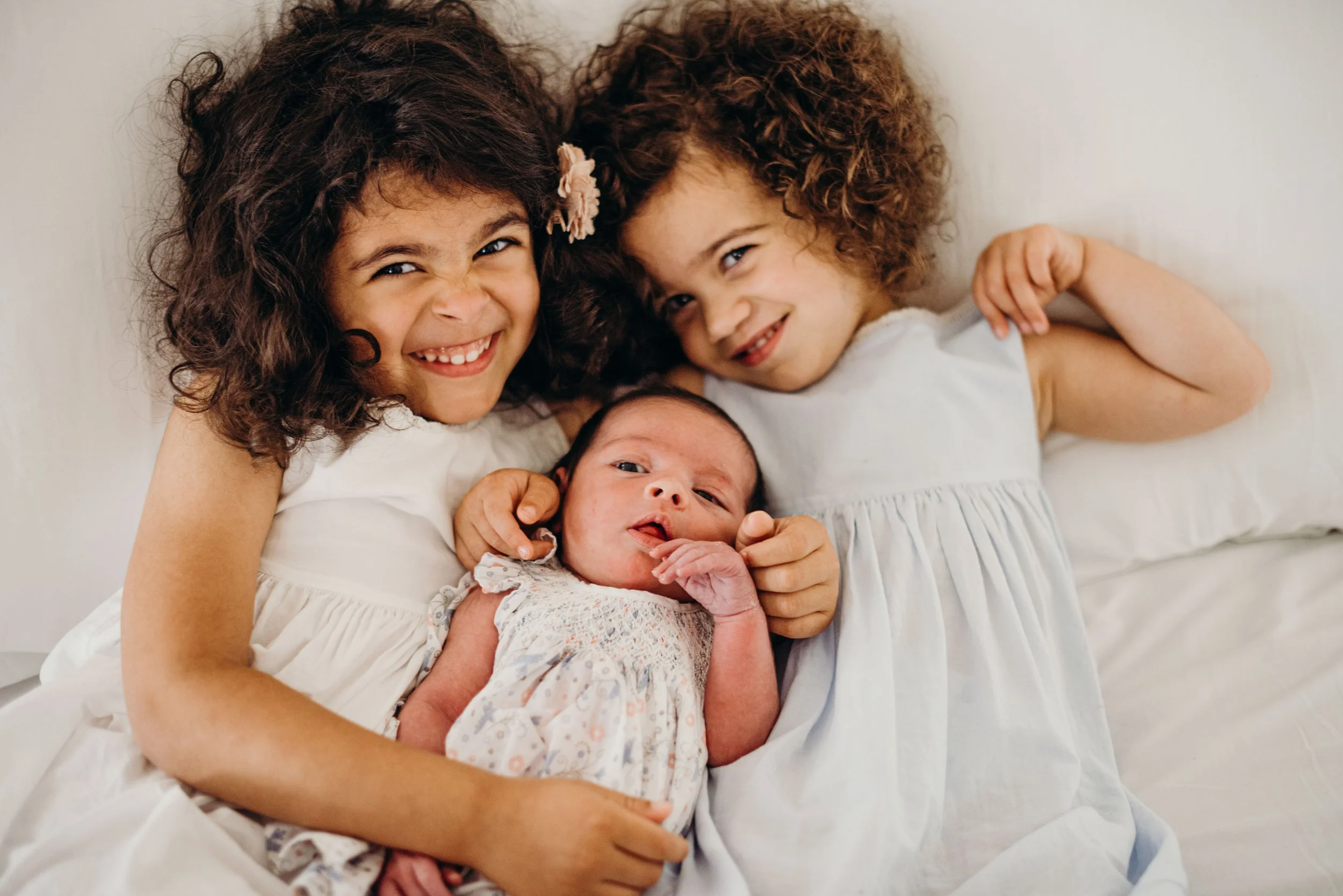 Three smiling children, two girls and a baby, lying on a bed with white sheets, posing for a photo. The girls have curly hair and are wearing white dresses, and the baby is also in a white dress with a floral pattern.