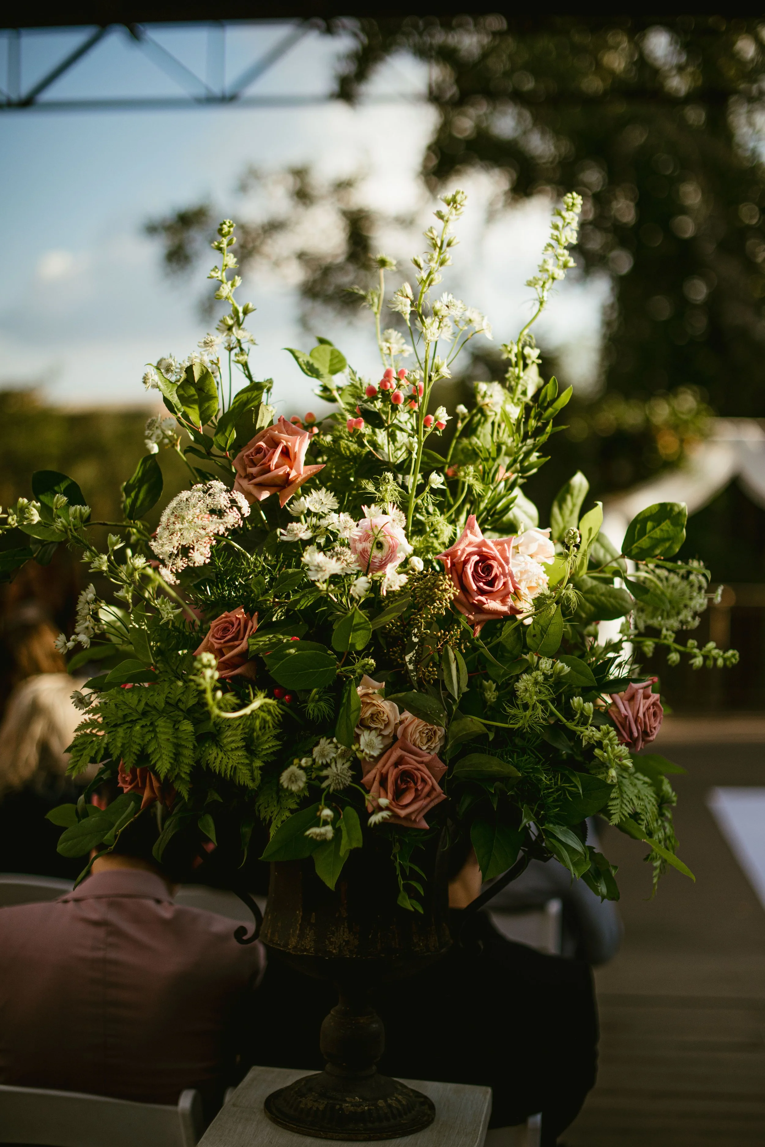 An elaborate floral arrangement in a vintage urn on a small table at an outdoor event, with people seated in the background.