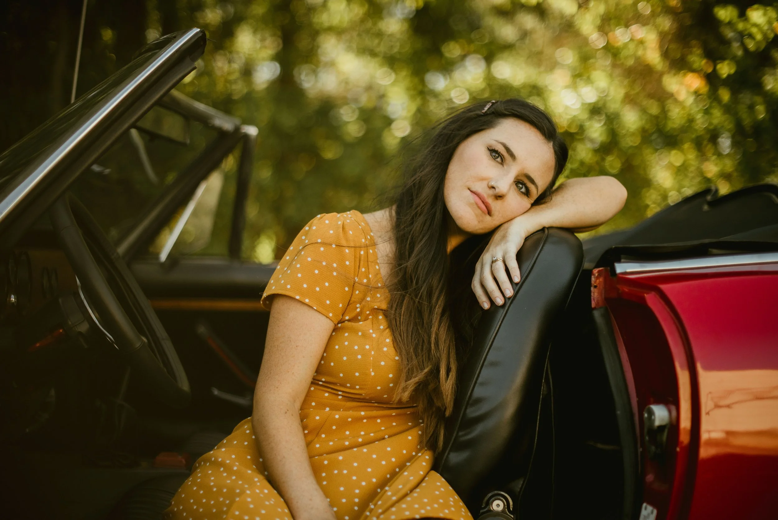 A young woman with long dark hair and freckles, resting her head on her arm, sitting inside a red convertible car during daytime with blurred green trees in the background. She is wearing a yellow dress with white polka dots.