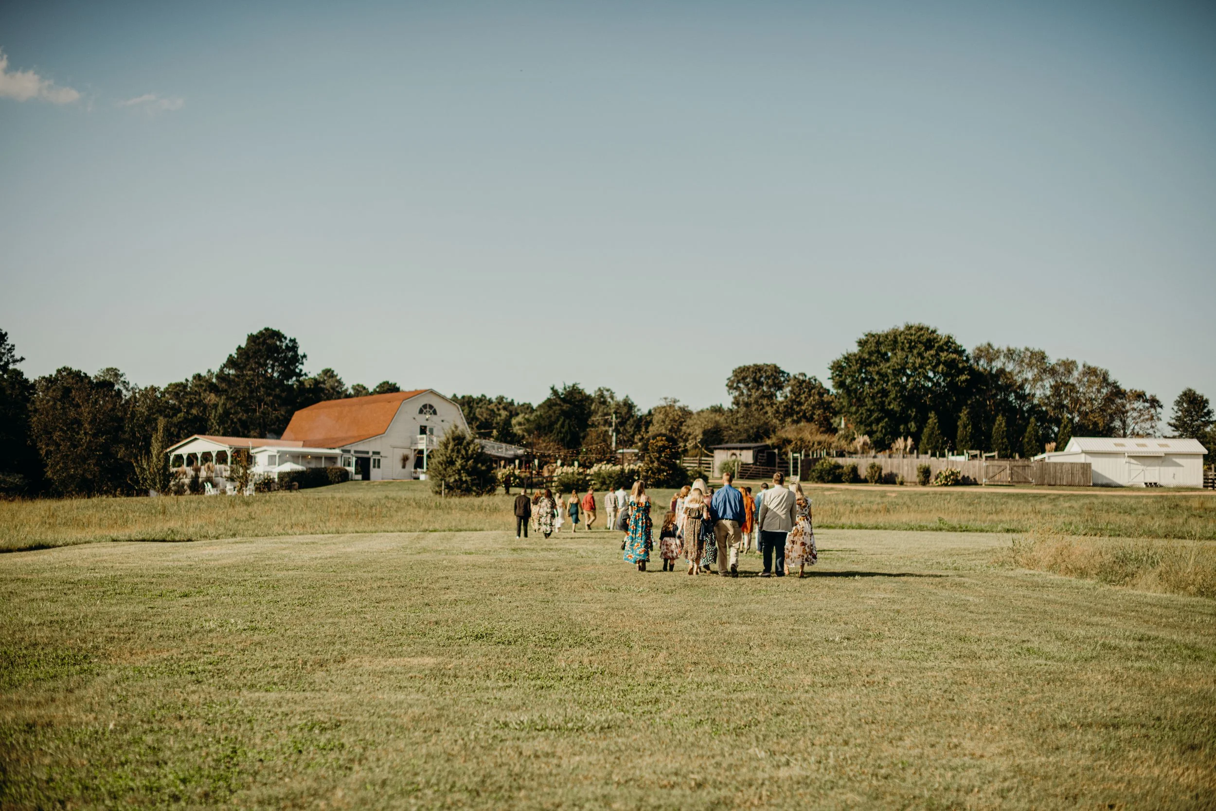 People walking across a grassy field towards houses and trees under a clear sky.