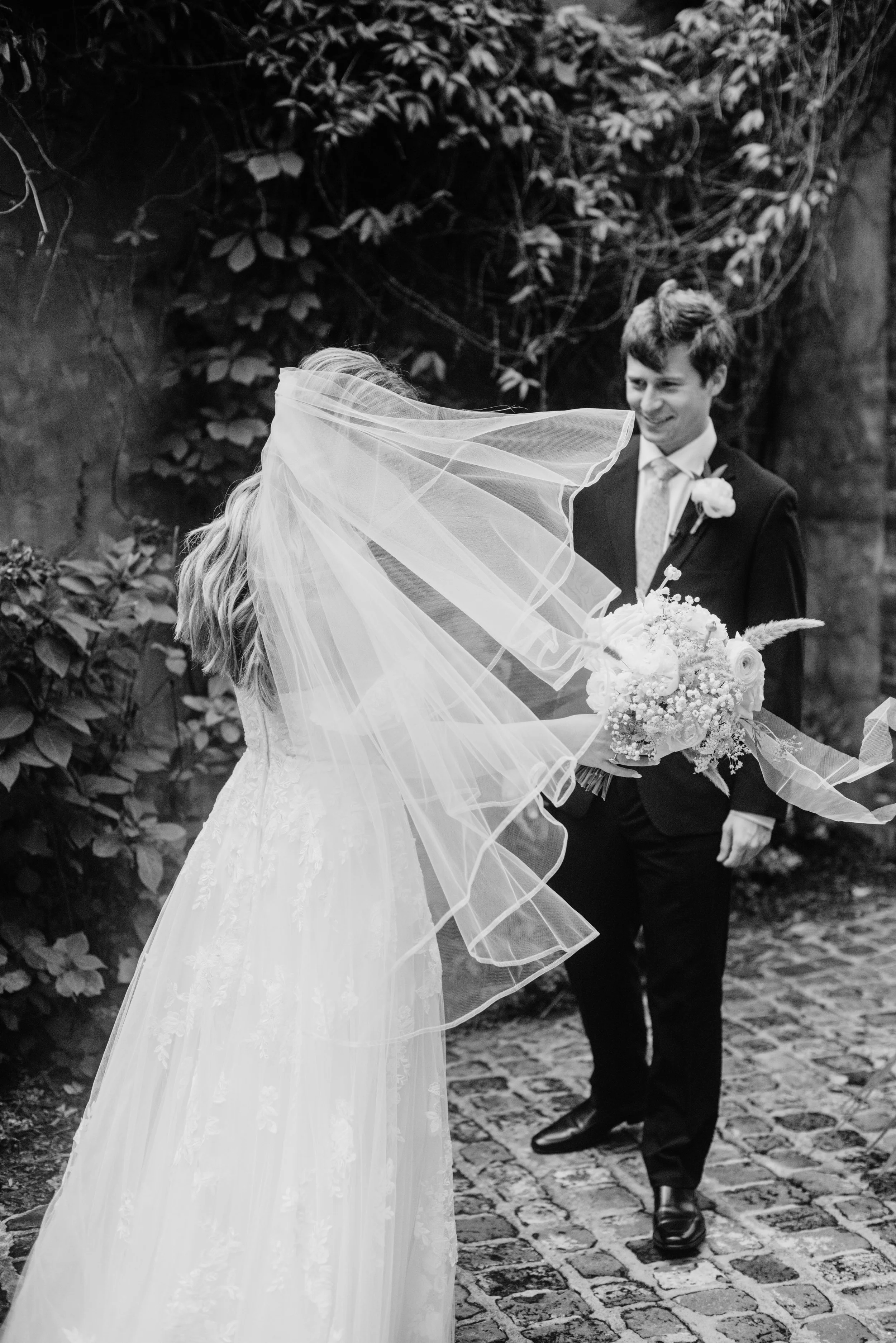 Black and white photograph of a bride and groom on their wedding day. The bride, in a lace wedding gown with a long veil, is holding a bouquet of flowers. The groom, dressed in a suit with a flower boutonniere, is smiling as he looks at her, standing