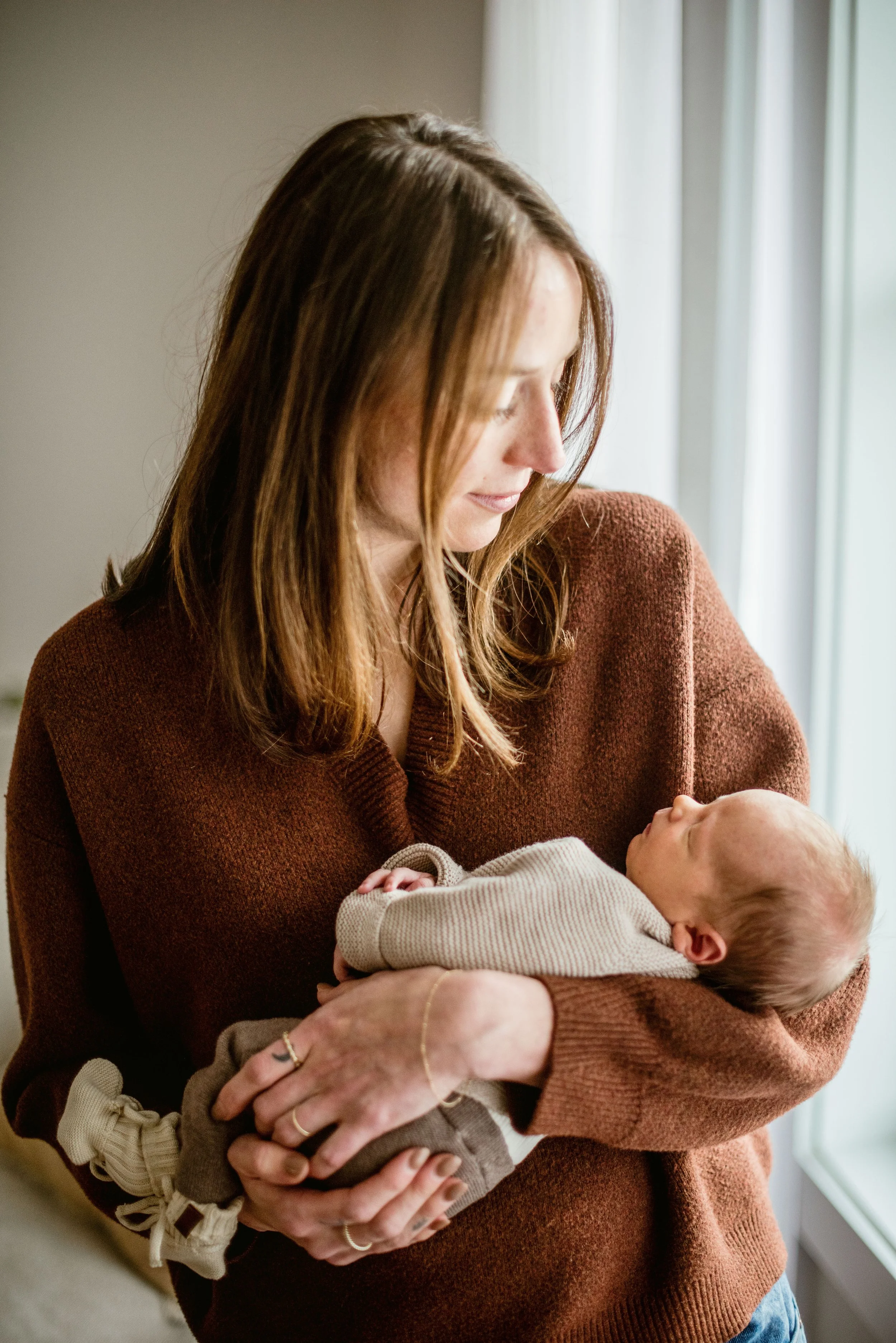 Woman in brown sweater holding a baby wrapped in a beige blanket, standing near a window with white curtains.