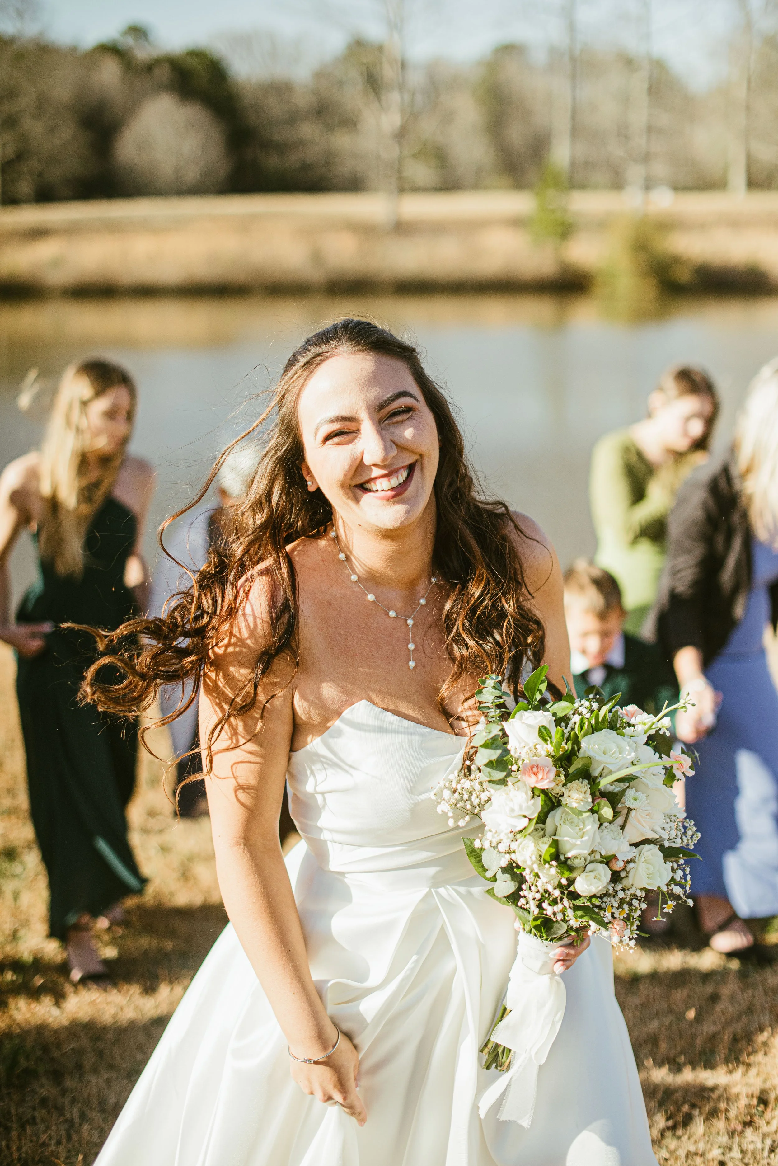 A smiling bride with long, wavy brown hair in a white wedding dress, holding a bouquet of white and pink flowers, outdoors by a body of water with trees in the background.