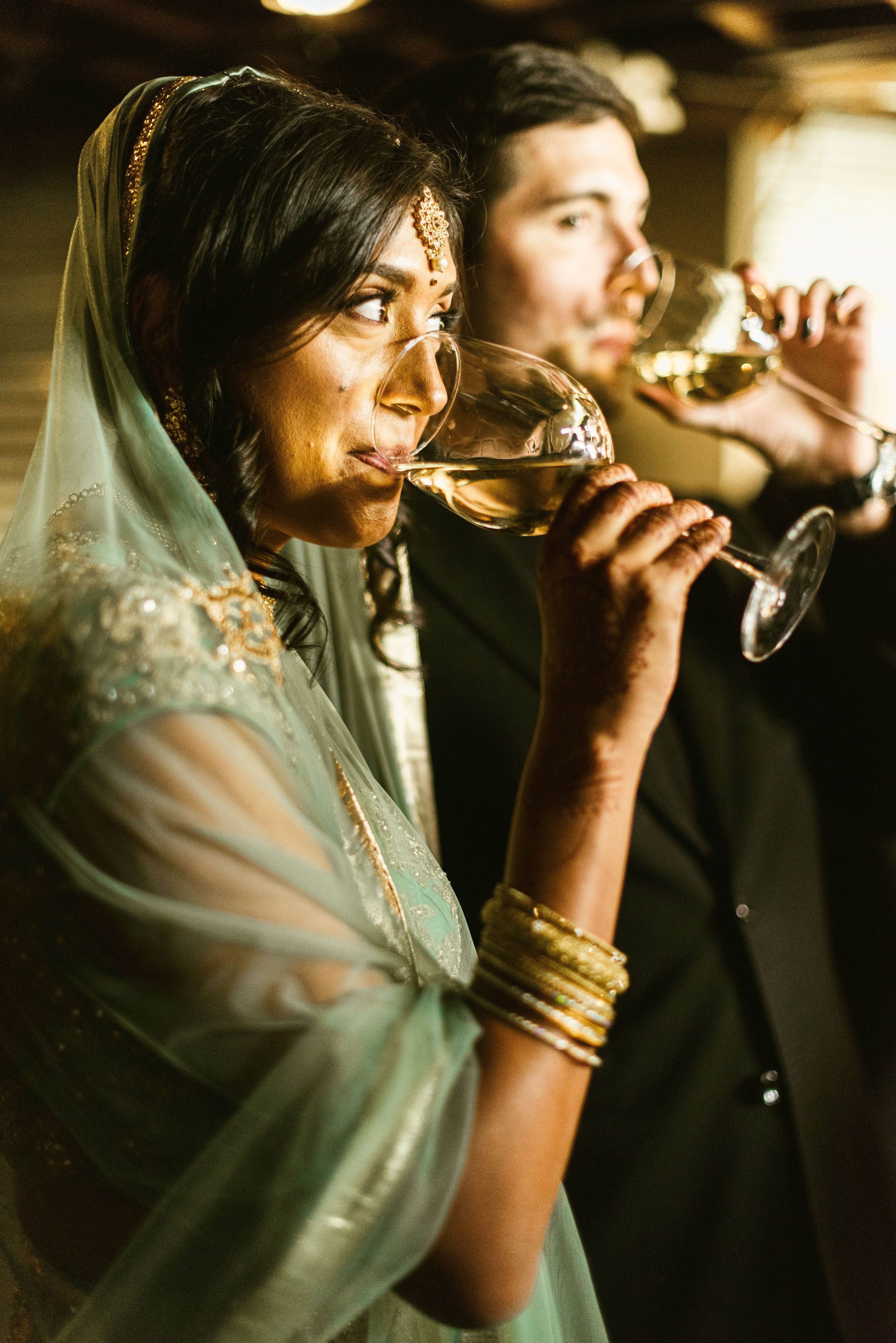 A woman in traditional Indian attire and jewelry, wearing a green sari, and glasses, drinking from a wine glass, with a man in a suit drinking from a similar glass in the background.