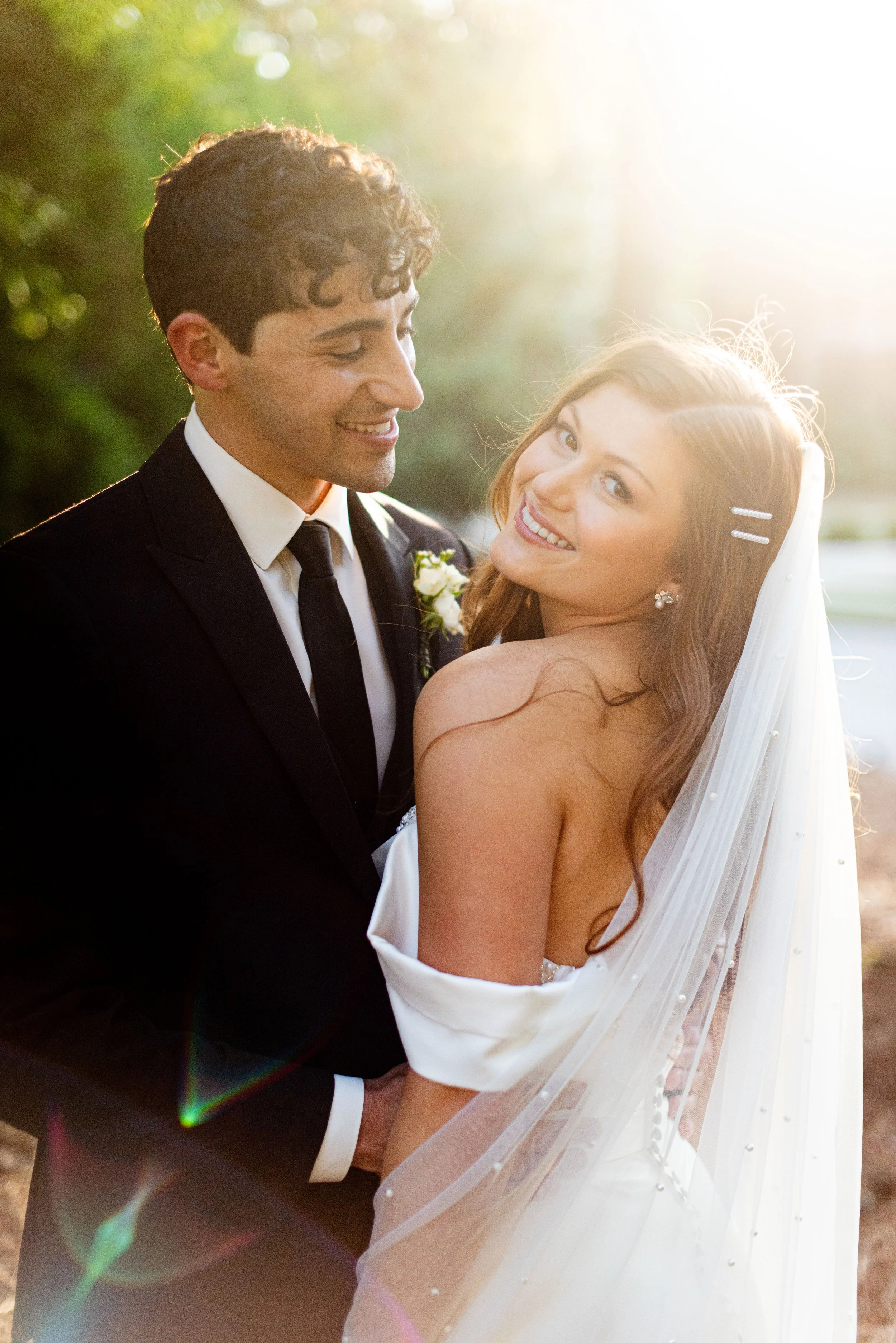 A smiling bride and groom on their wedding day, with the groom wearing a black tuxedo and the bride in a white wedding dress with a veil. The sun shines brightly behind them, creating a warm glow.