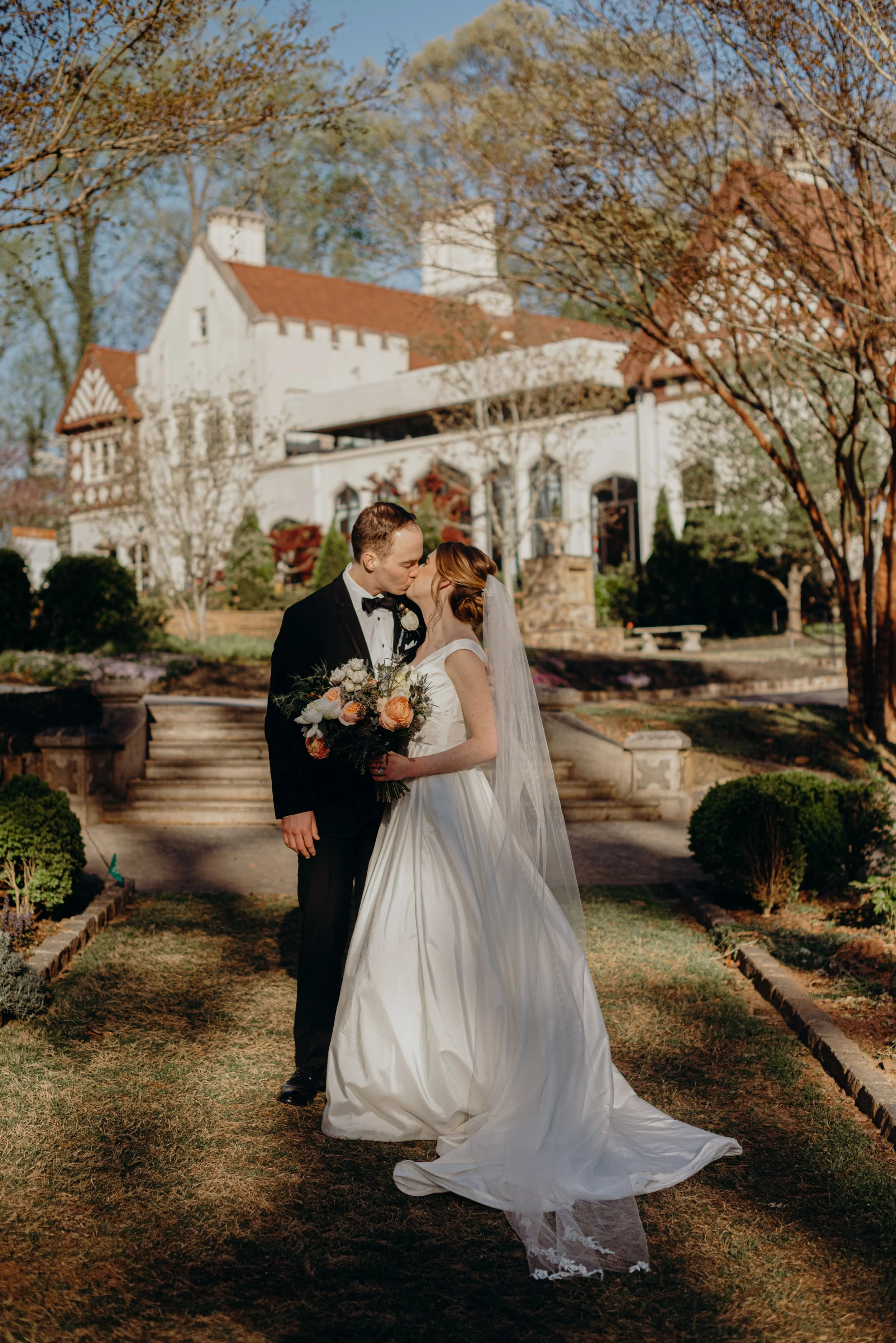 A bride and groom kissing outdoors in front of a white, castle-like house during daytime, with trees displaying autumn foliage.