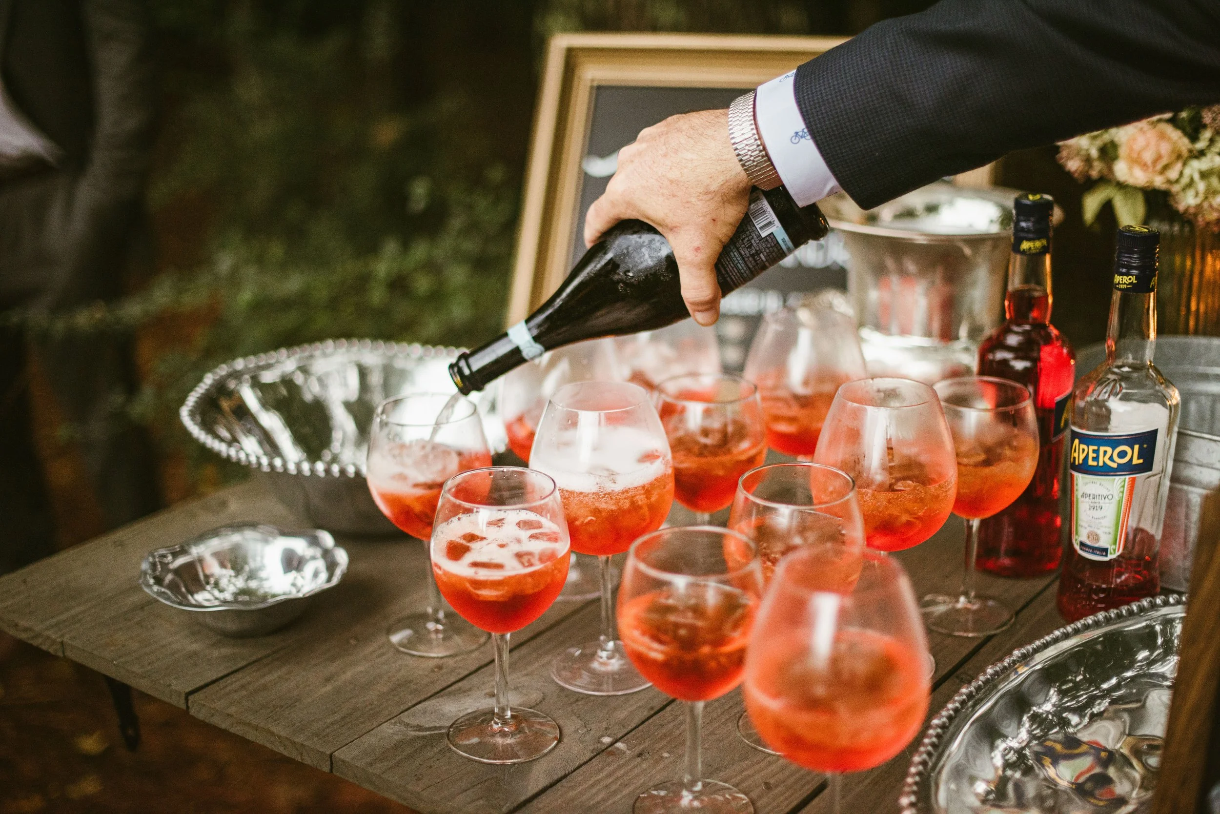 A person in a dark suit and watch pouring pinkish-orange beverage from a dark bottle into glasses at an outdoor event. There are multiple glasses, some filled with ice and the drink, on a wooden table with decorative silver trays and bottles of Apero